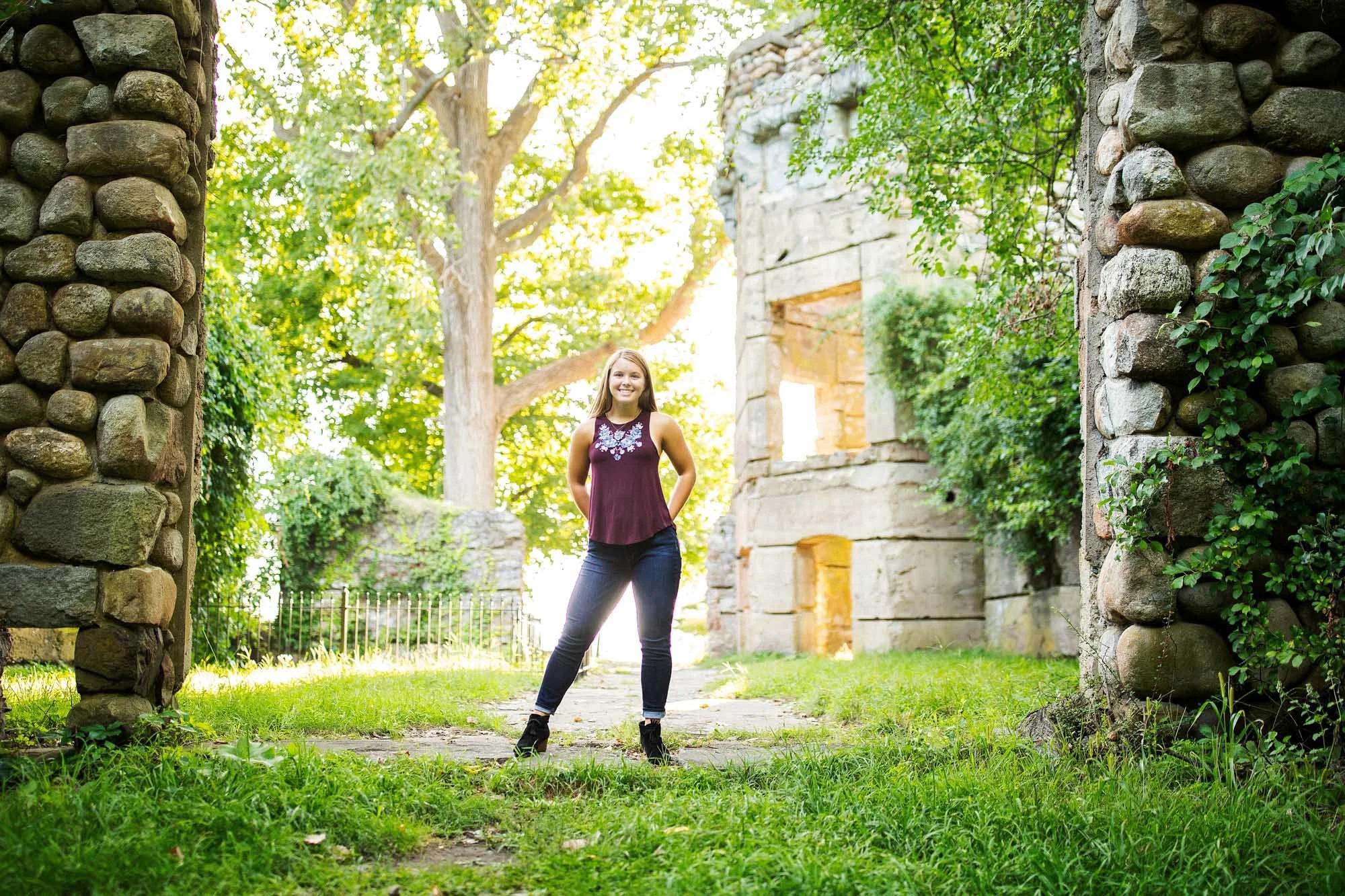 A smiling woman stands outdoors between stone ruins, surrounded by green trees and grass on a sunny day.