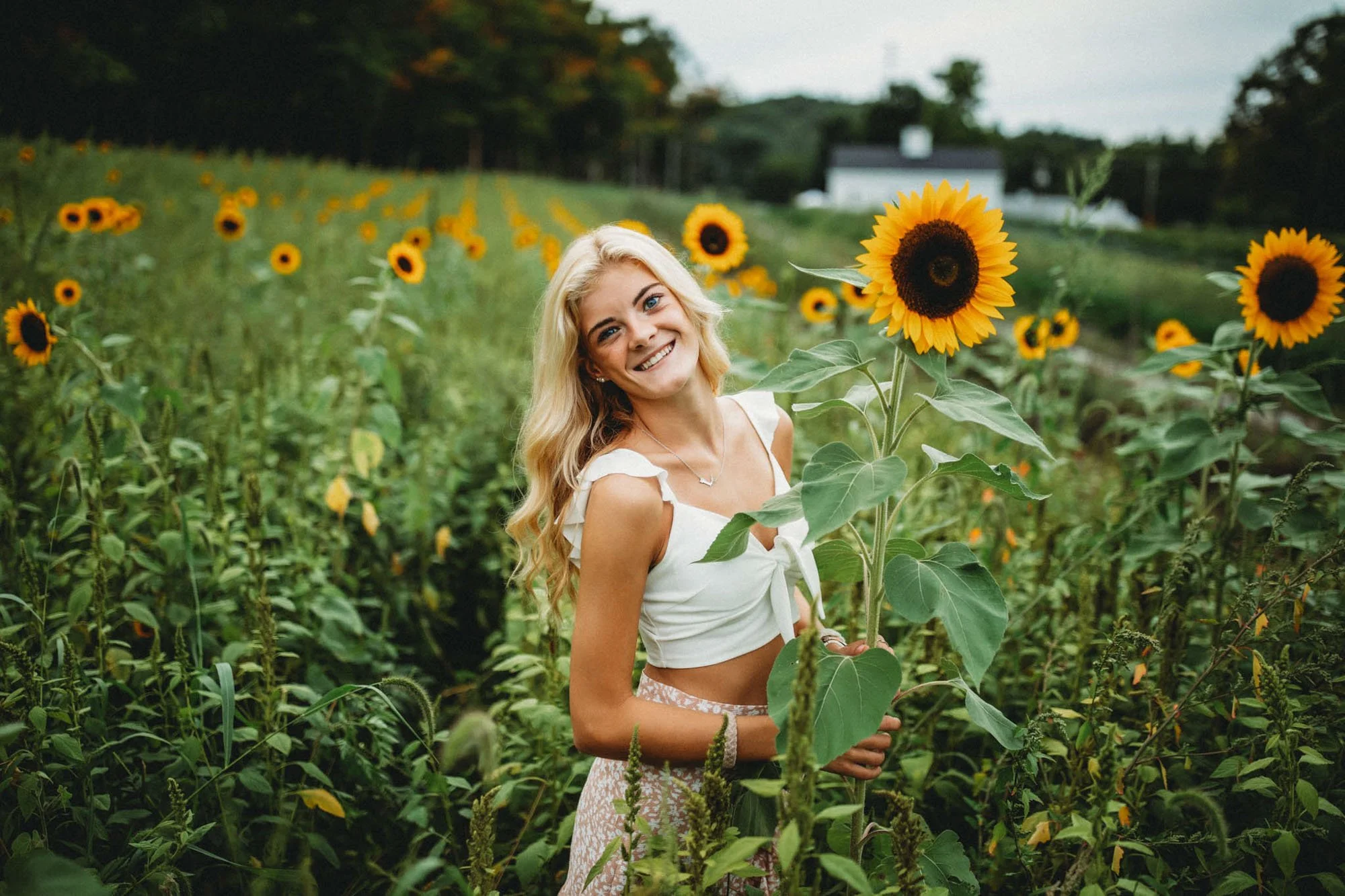 A smiling young woman with long blonde hair standing in a sunflower field during daytime, wearing a white sleeveless top and patterned skirt.