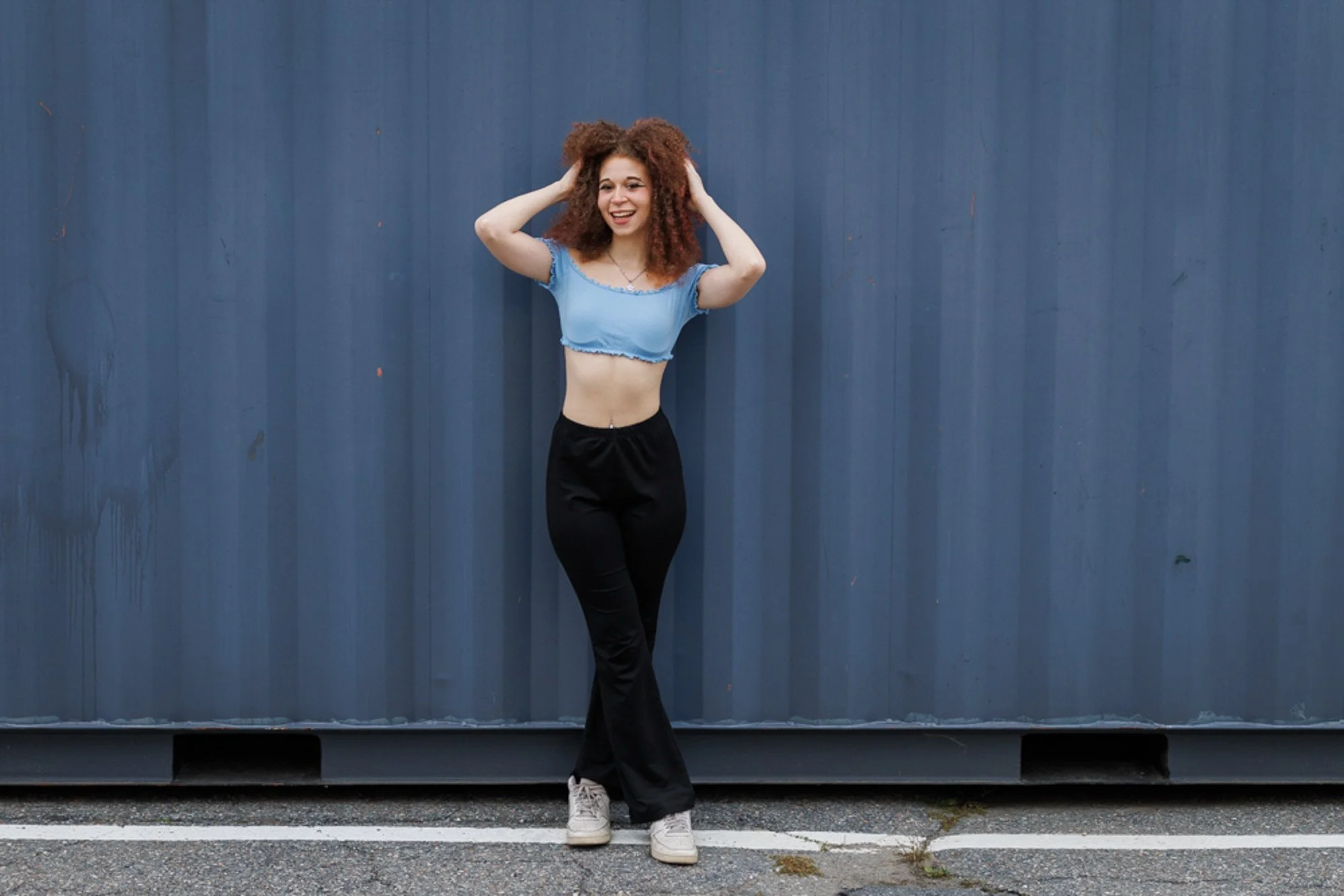 Young woman with curly red hair standing in front of a blue metal wall, wearing a cropped blue t-shirt, black pants, and white sneakers, smiling with hands in hair.