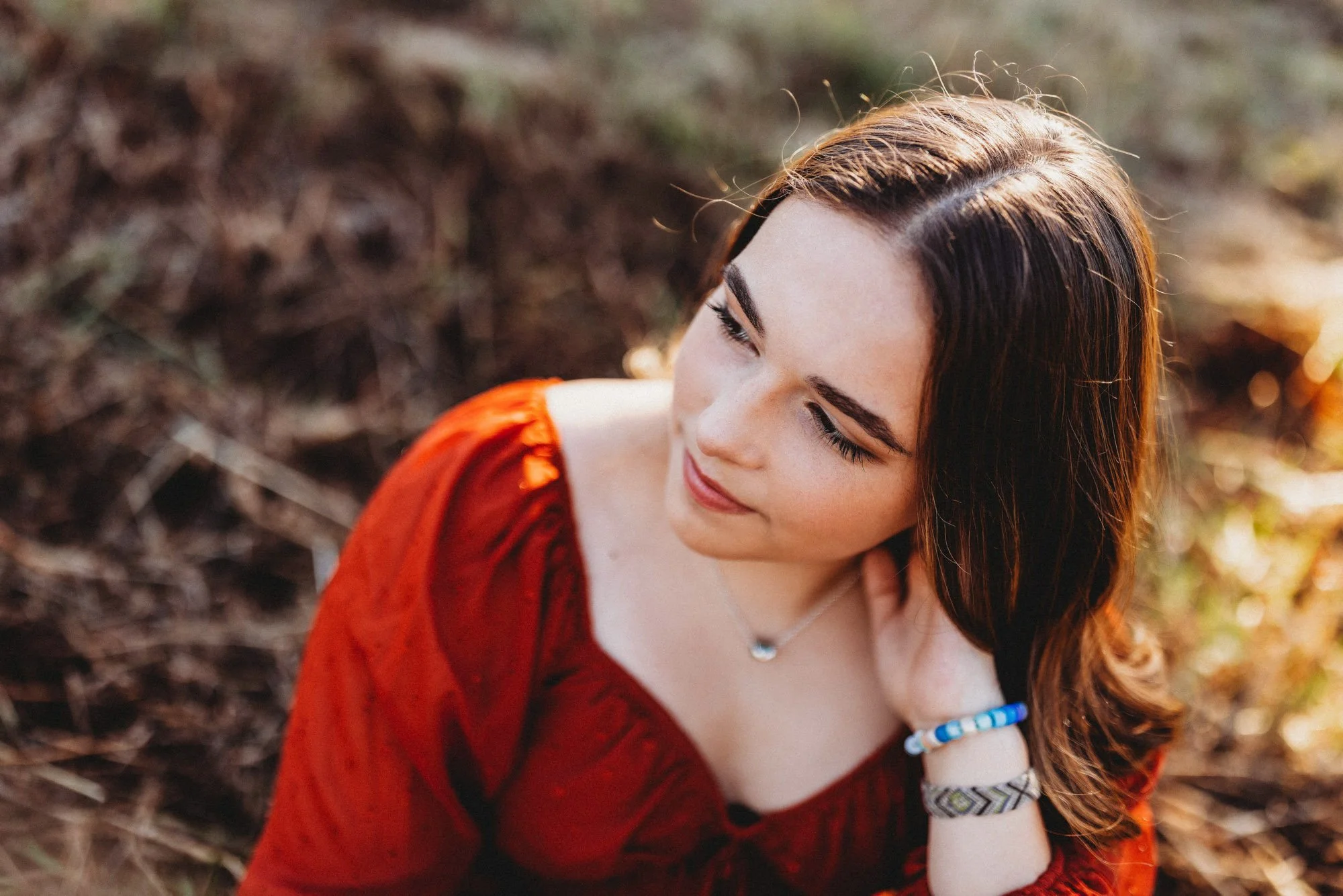 A young woman in a red dress with brown hair looking down and smiling outdoors during sunset.