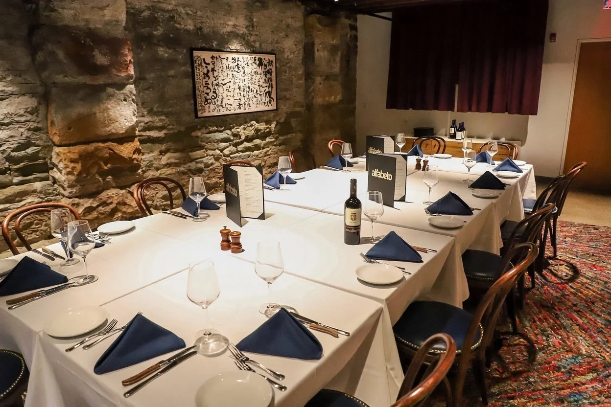 A restaurant dining table set for a meal with wine glasses, napkins, plates, and silverware, against a stone wall with a framed artwork, and a bottle of wine on the table.