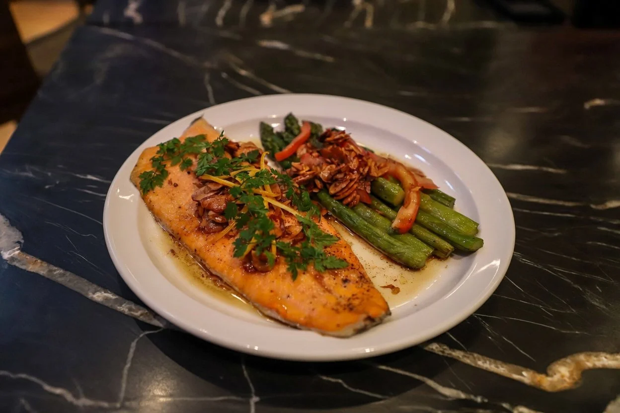 A plate of cooked salmon topped with herbs and vegetables, served with asparagus and a vegetable medley, on a black marble table.