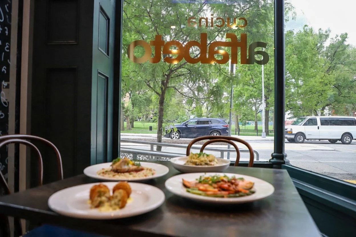 Four plates of food on a table inside a restaurant near a window with trees and cars outside.