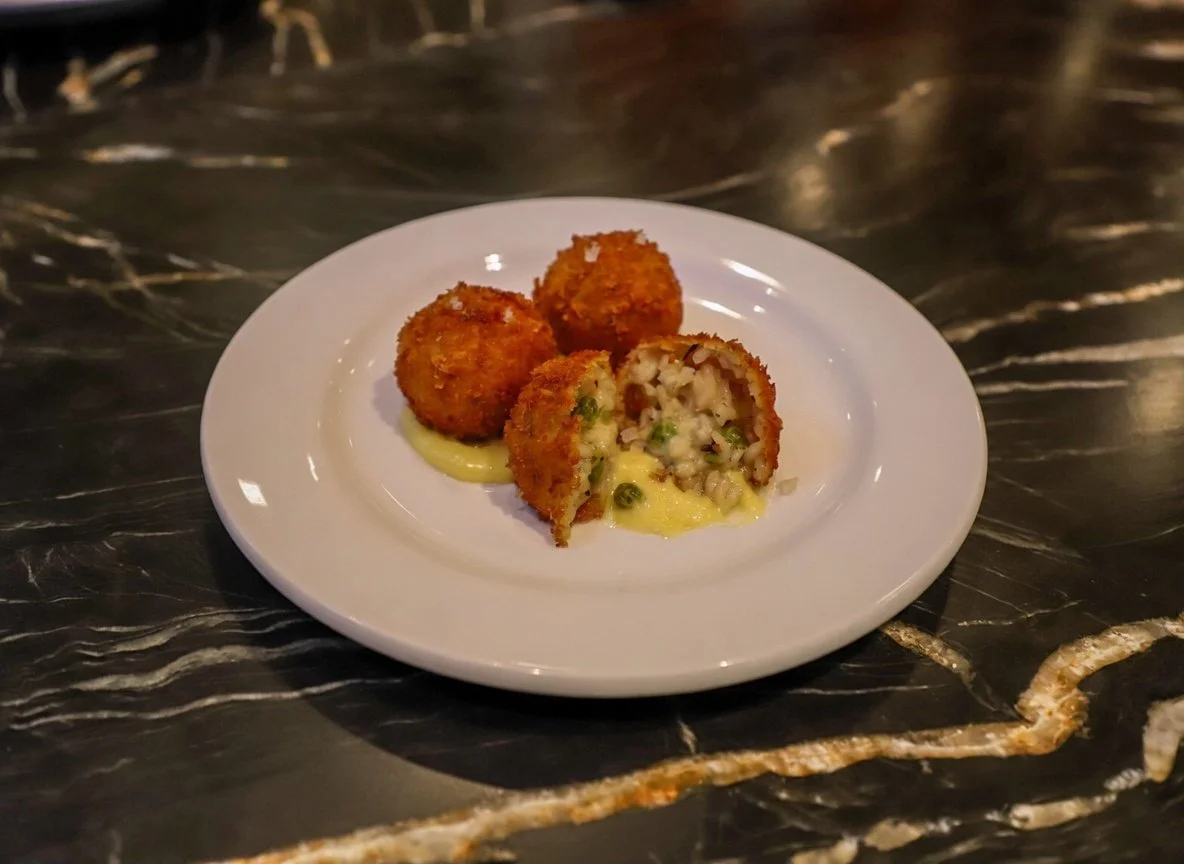 Three fried croquettes on a white plate, one cut in half to reveal creamy filling with green peas and other ingredients, all on a dark marbled surface.