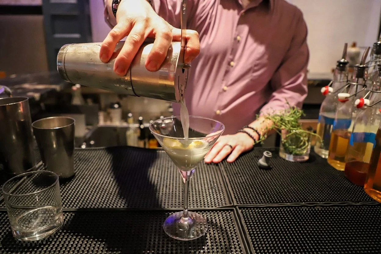 Person pouring a cocktail into a martini glass at a bar counter.