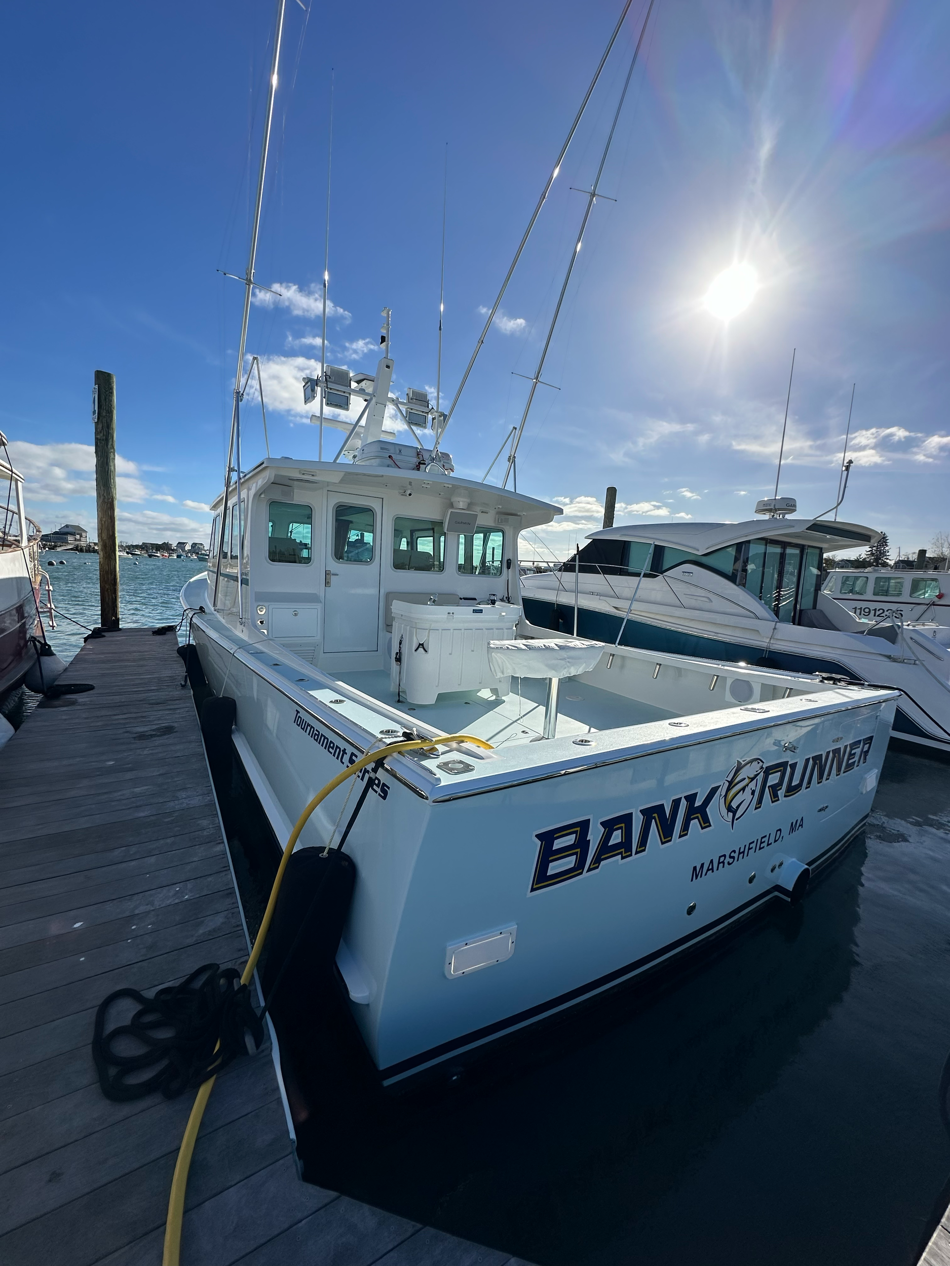 A white boat named 'Bank Runner' docked at a marina in Marshfield, MA, with other boats visible in the background and the sun shining brightly in the clear blue sky.