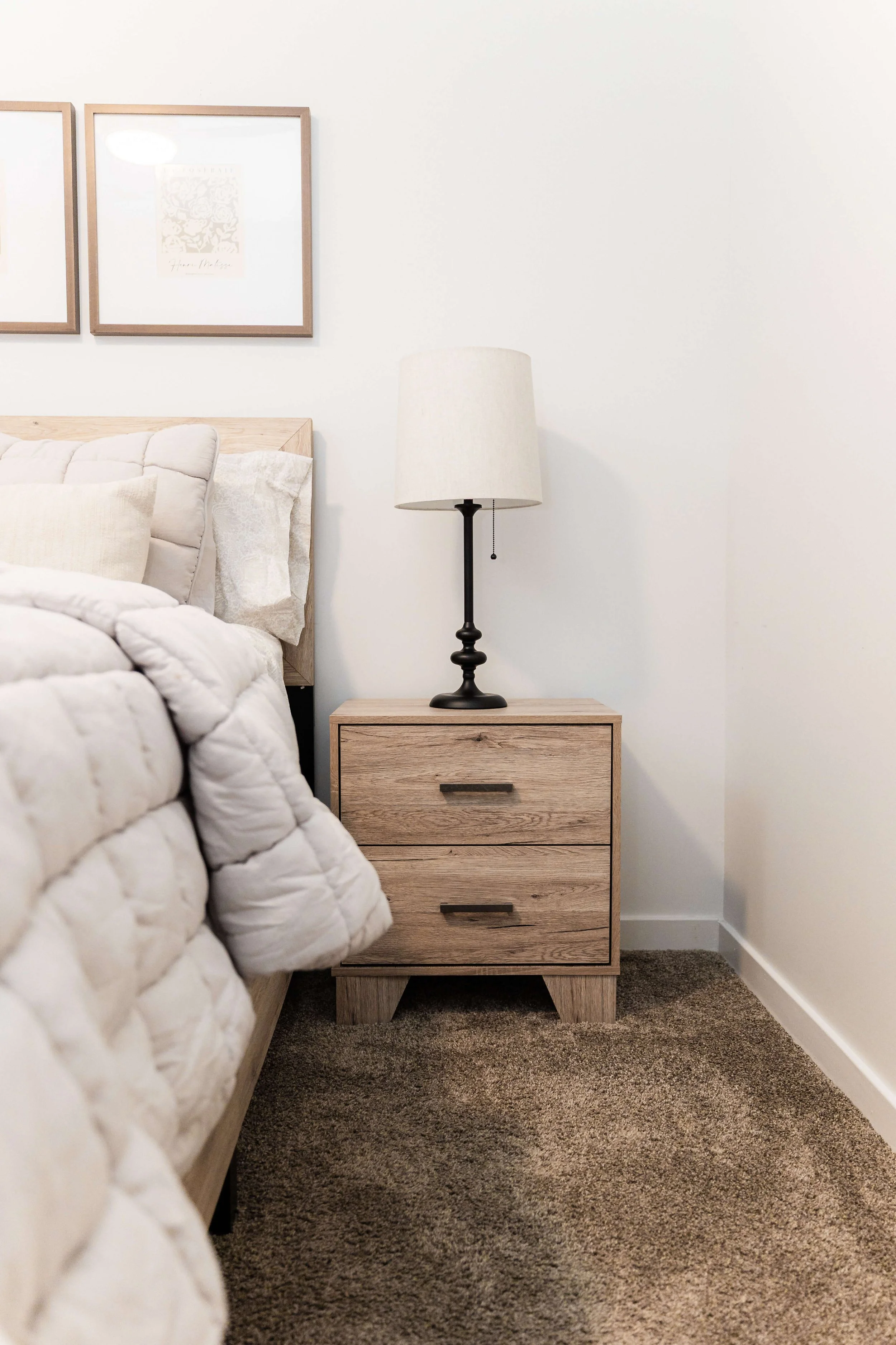 A quiet, hotel-inspired bedroom moment — a bedside lamp framed by soft bedding and coastal artwork.
