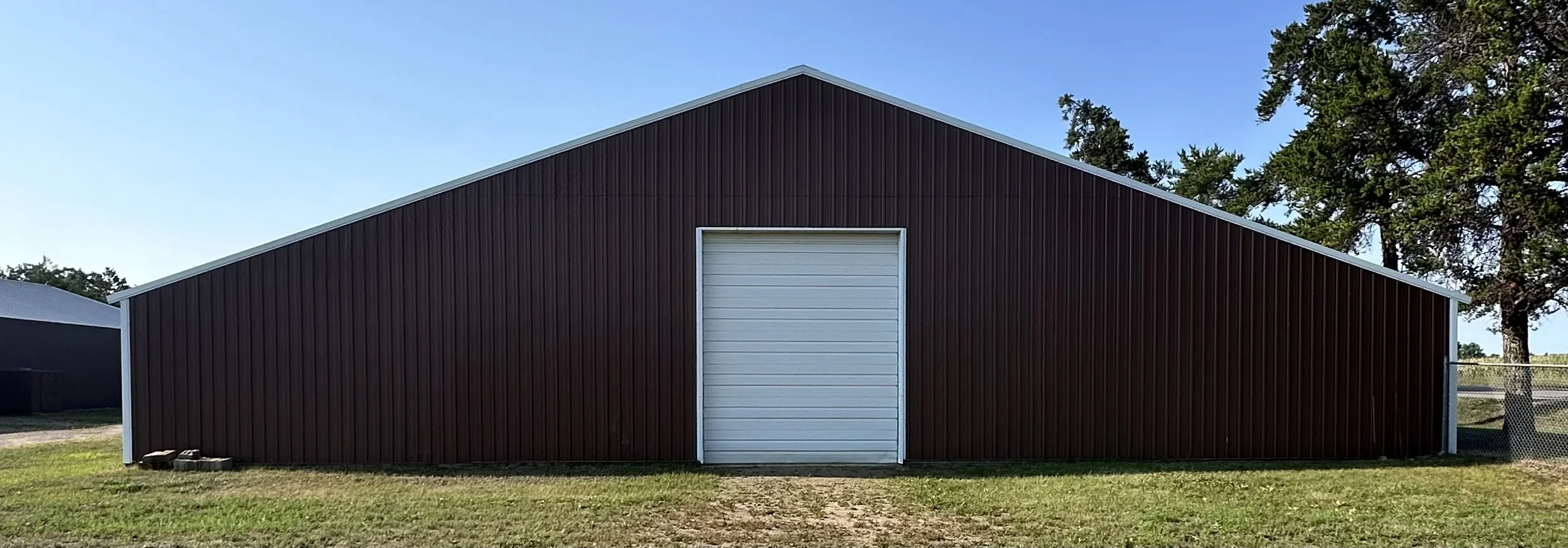A large brown metal storage building with a white roll-up door in the center, situated on a grassy area with trees and a clear blue sky background.