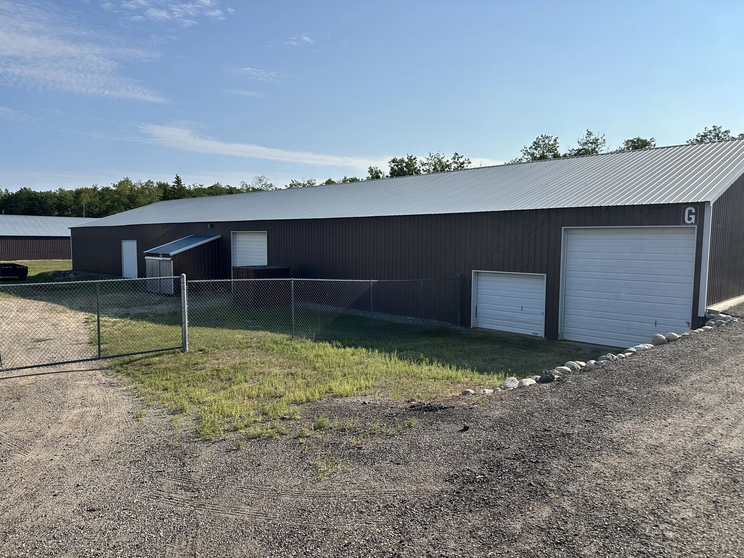 A large metal building with the letter G on the corner, a sliding garage door, a smaller door, and a sloped roof, surrounded by a gravel and grass yard with a chain-link fence, under a clear blue sky with some clouds.