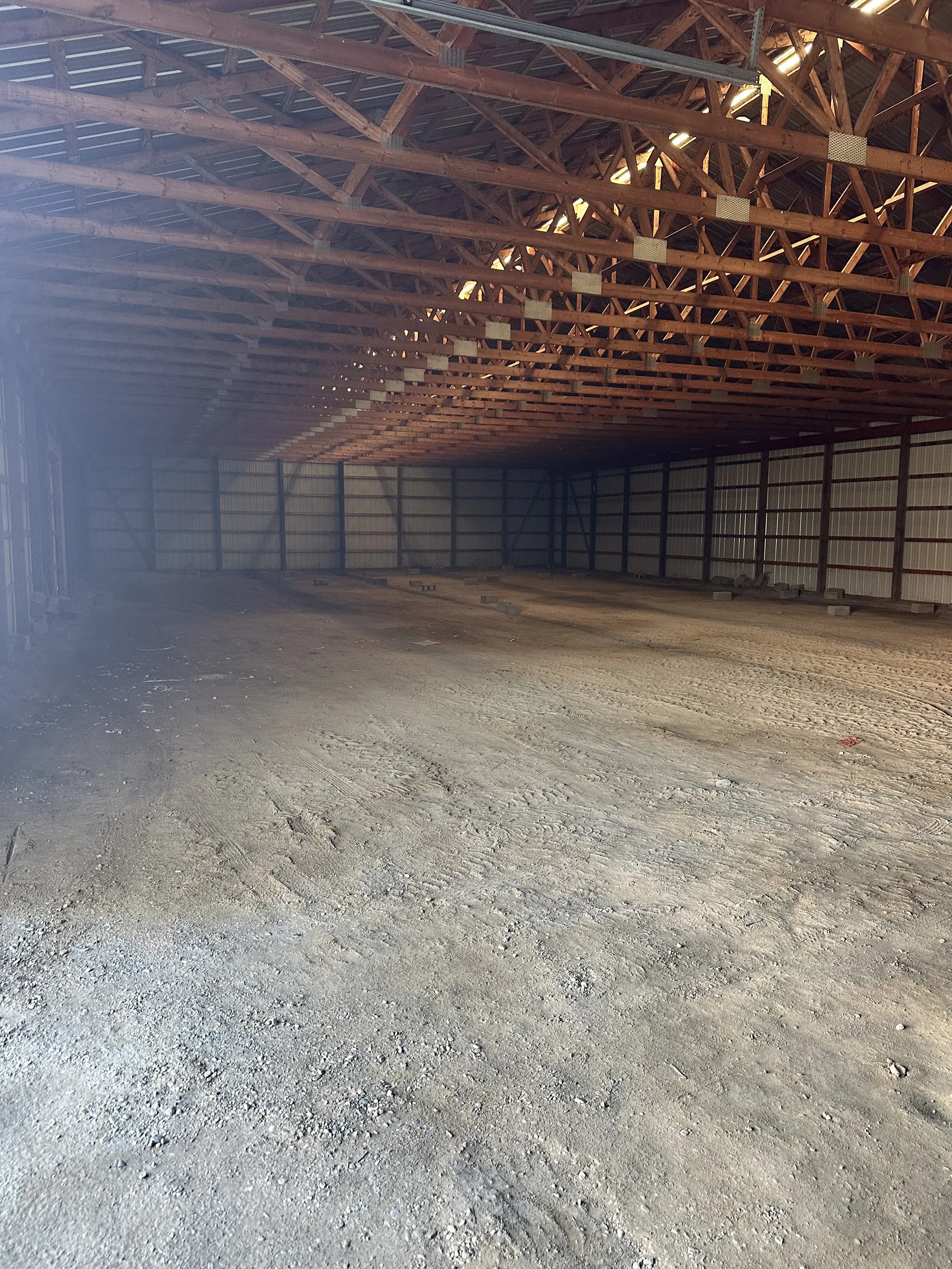 The interior of a large metal building under construction, showing exposed wooden roof trusses and a dirt floor.