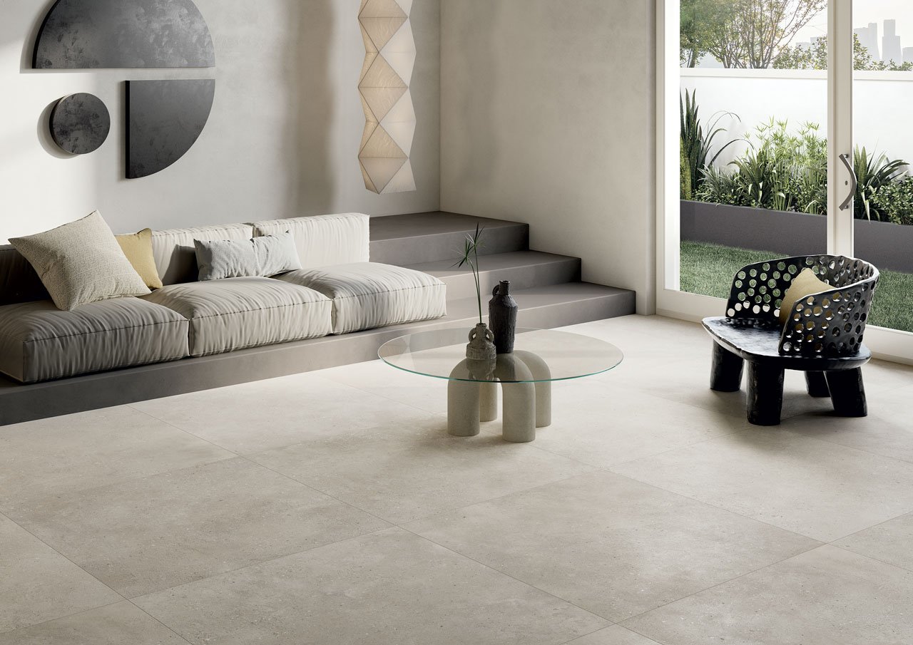 Modern living room with a beige sofa, decorative pillows, a glass coffee table with vases, a black sculptural chair, sliding glass doors leading to a garden, and minimalist wall art.