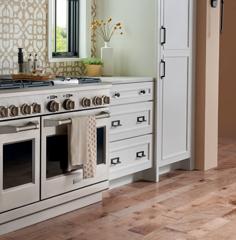 Kitchen with white cabinets, a white oven with beige knobs, a stovetop, a towel hanging on the oven handle, a window, decorative plants and objects on the countertop, and natural wood flooring. Atlas Homewares