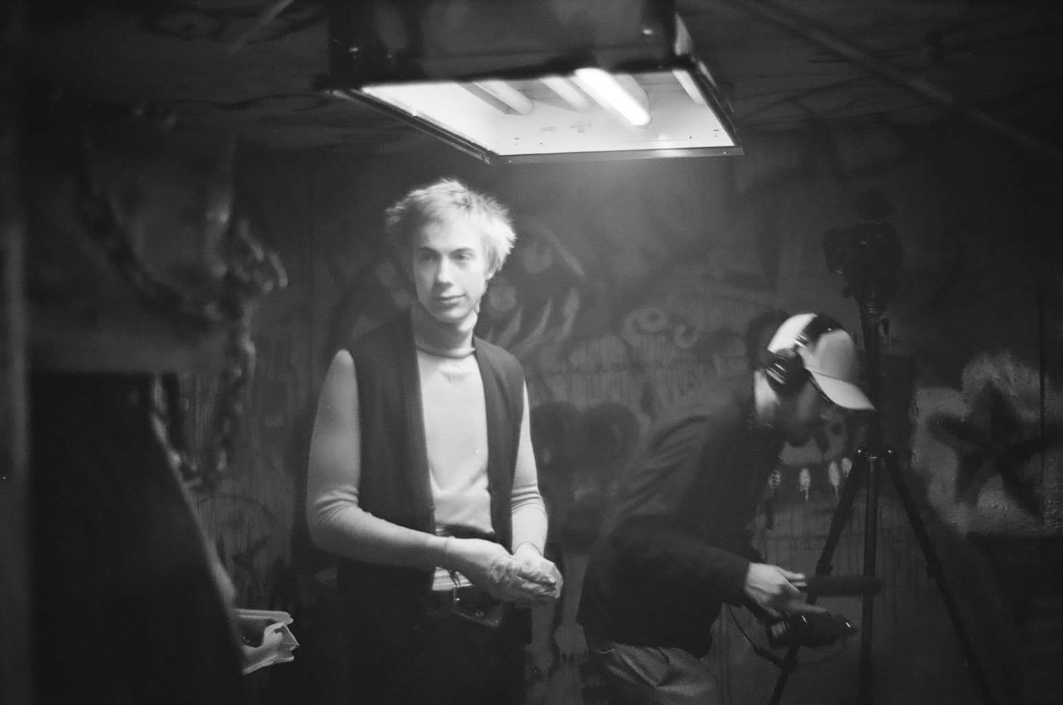 A black and white photo of a young man with messy hair and casual clothing, standing in a room with graffiti-covered walls, with a person in a cap and headphones working with equipment nearby.