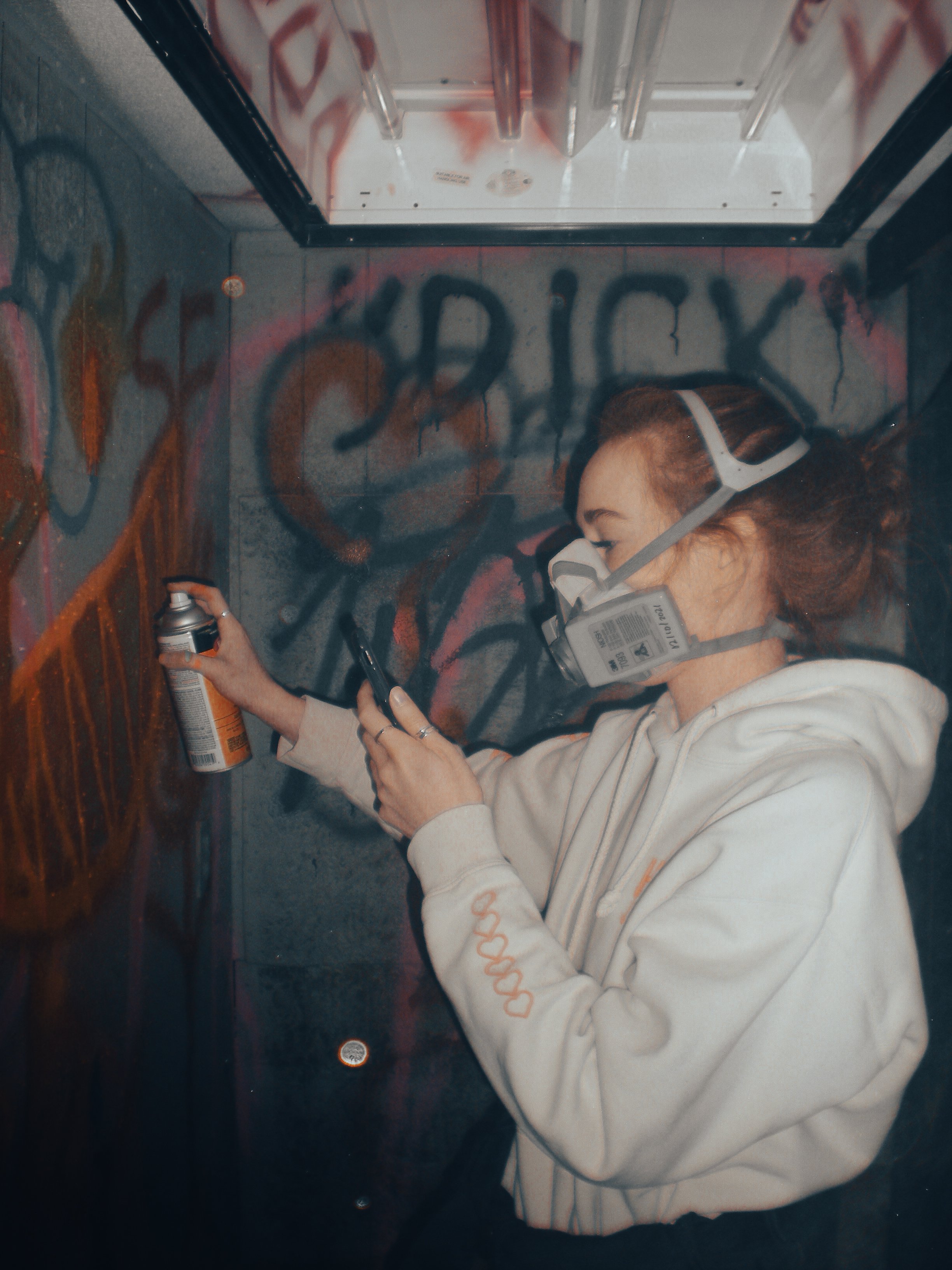 A young woman wearing a respirator mask and hoodie spray paints graffiti on a wall.