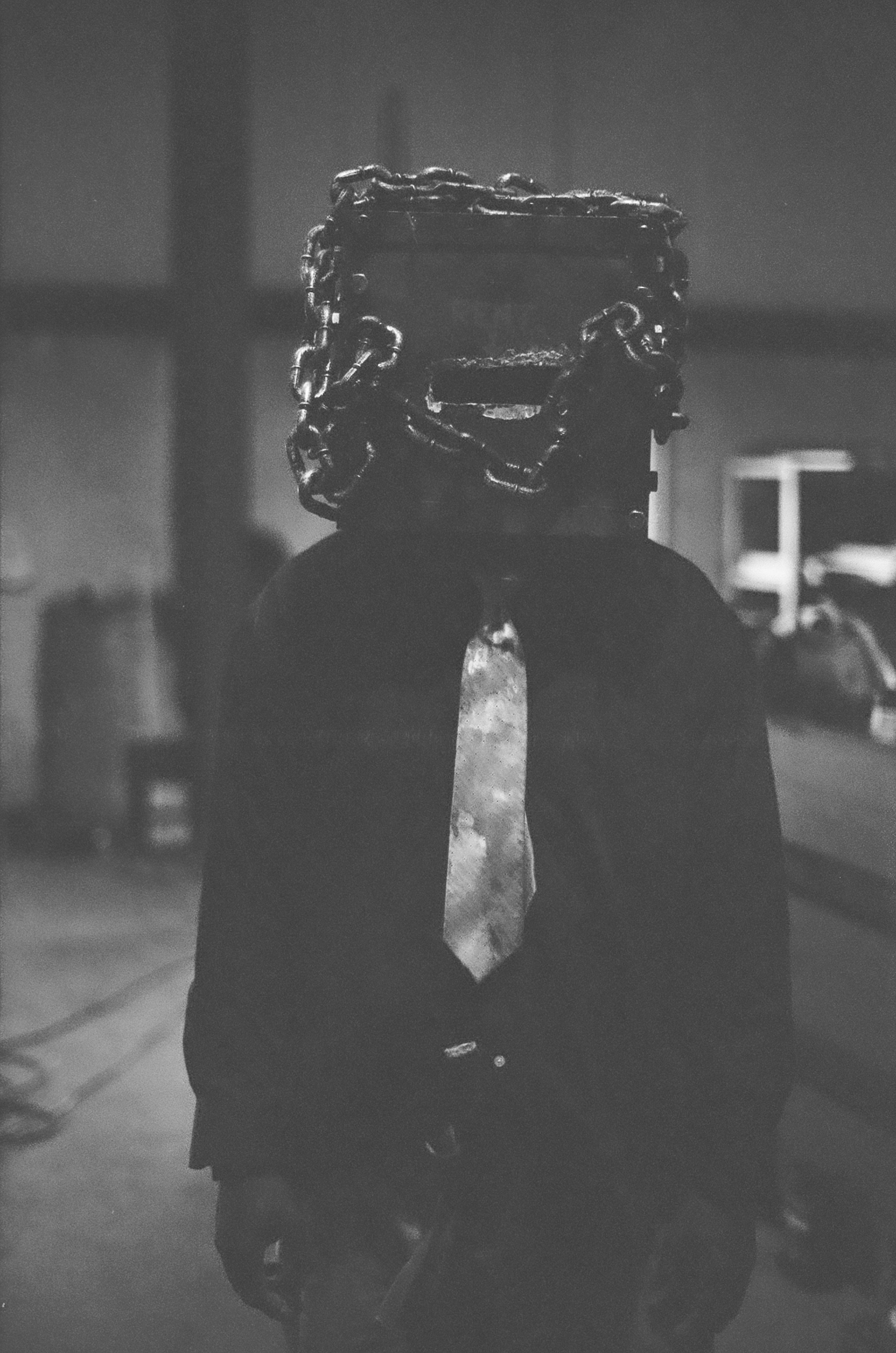 A person dressed in business attire with a shirt and tie, but their head is replaced by a metal container welded with chains, creating a surreal, artistic image.