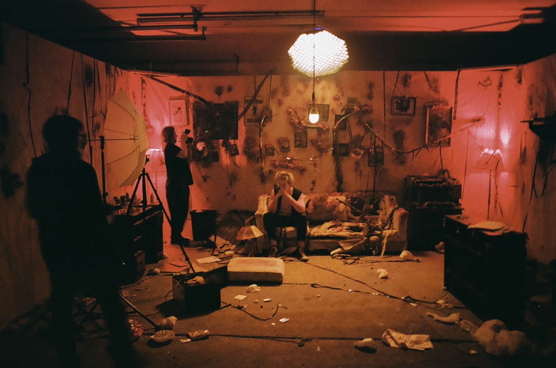 A person sitting on a chair in a messy, dimly lit room with scattered objects, photographs, and string decorations on the wall, illuminated by a hanging light bulb and a recent pinkish-orange light.