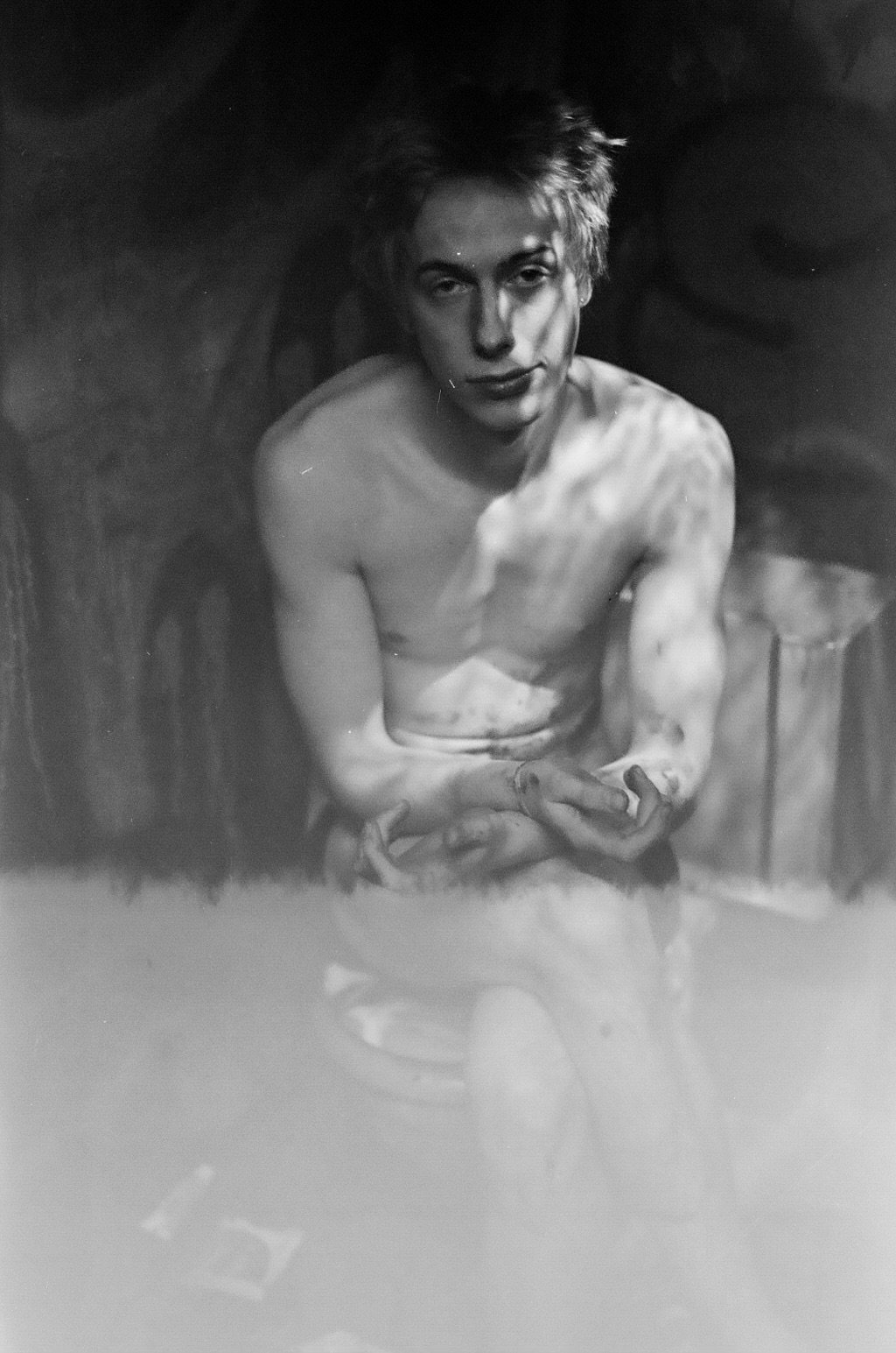 Black and white photo of a shirtless young man with short, messy hair looking at the camera. He is sitting with his hands clasped and a serious expression on his face.