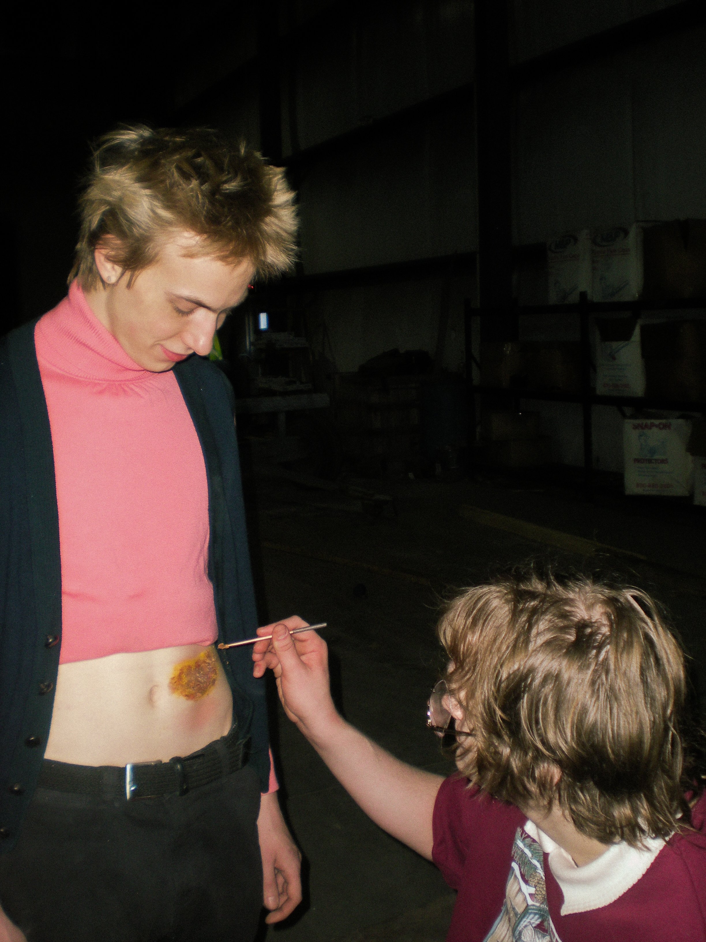A young woman with short, blonde hair getting her stomach painted with a design by a child with shoulder-length, light-brown hair wearing glasses. They are indoors, in a dimly lit space with shelves in the background.