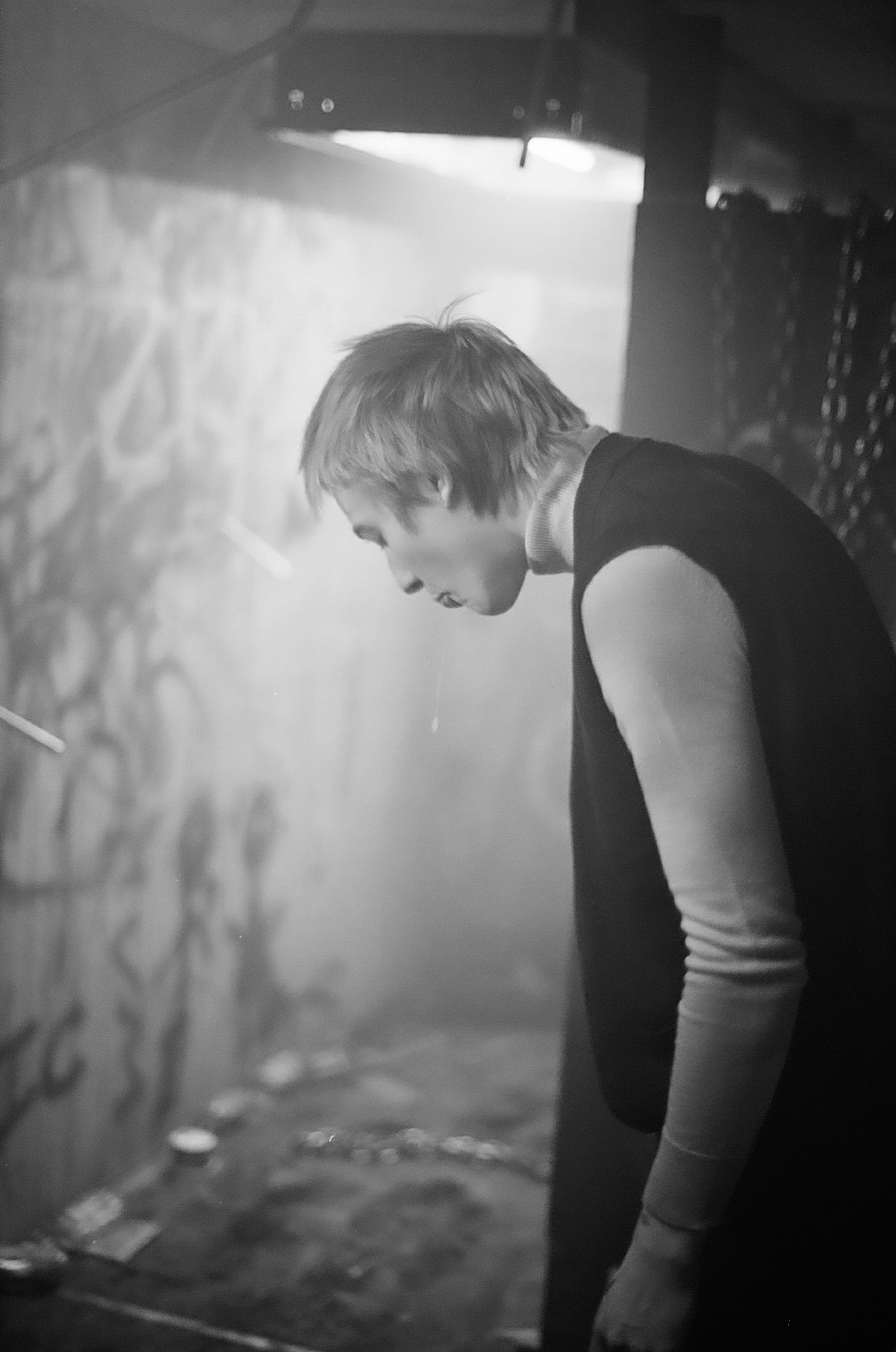 A young person with short hair, wearing a sleeveless shirt over a long-sleeve turtleneck, is standing on a skateboarding ramp, looking downward with a serious expression in a dimly lit indoor skate park.