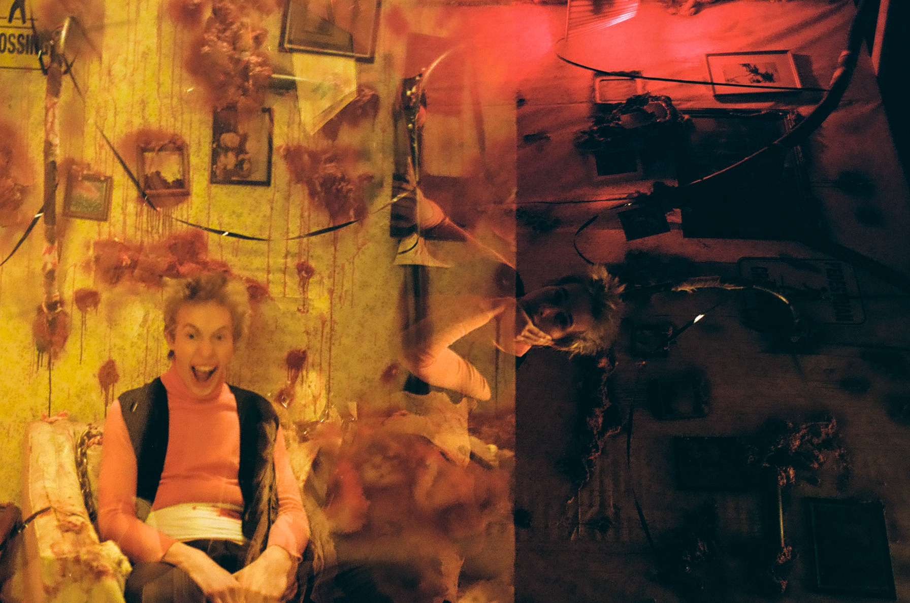 A double exposure photograph showing a woman with curly hair smiling widely and sitting on a vintage couch, with a wall decorated with framed pictures and a string of marionettes behind her, and a person with a dog, possibly taking a selfie, reflecte