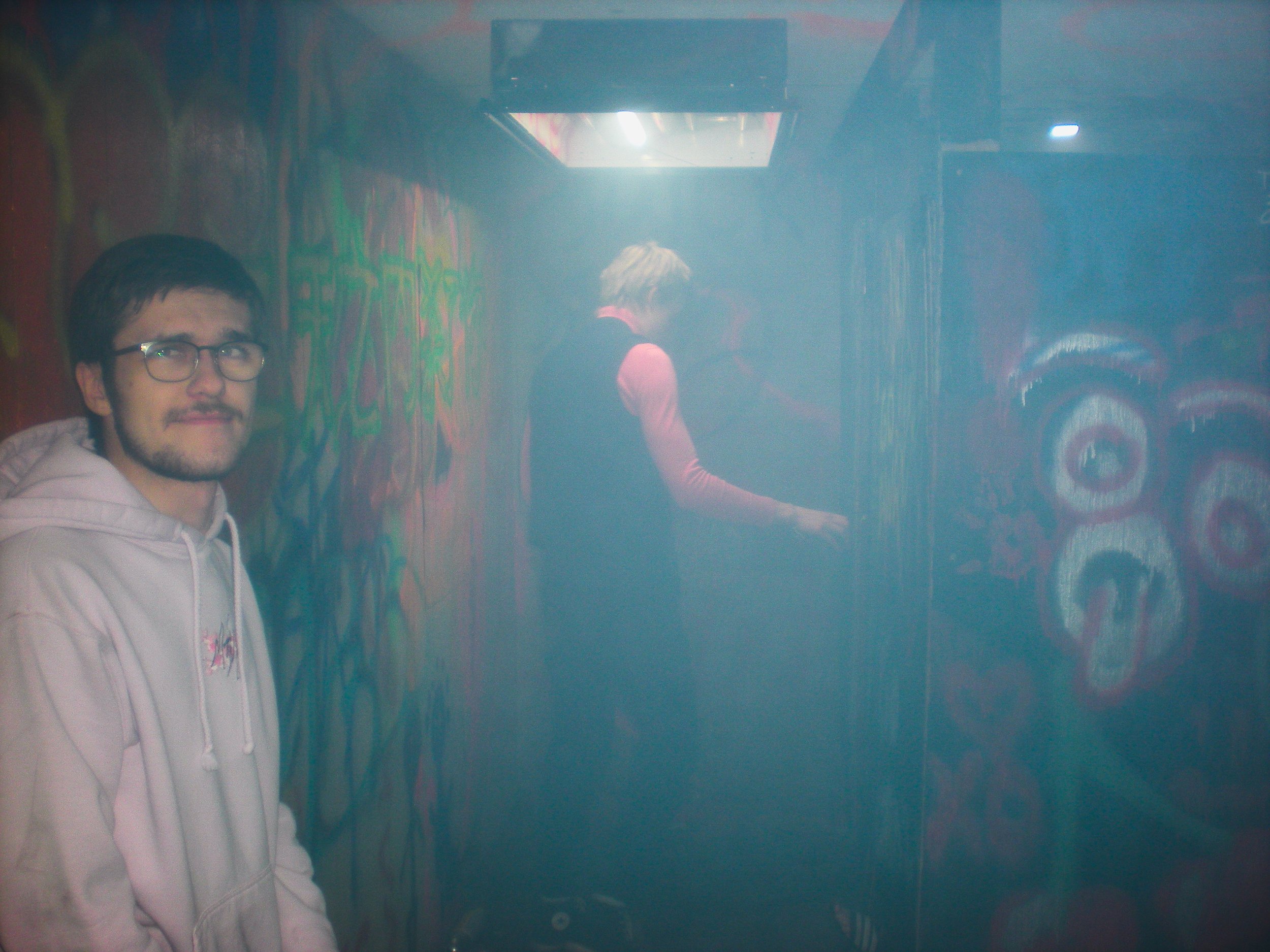 Young man with glasses and a beige hoodie standing in front of a foggy, colorful graffiti wall with a woman in the background near a door.
