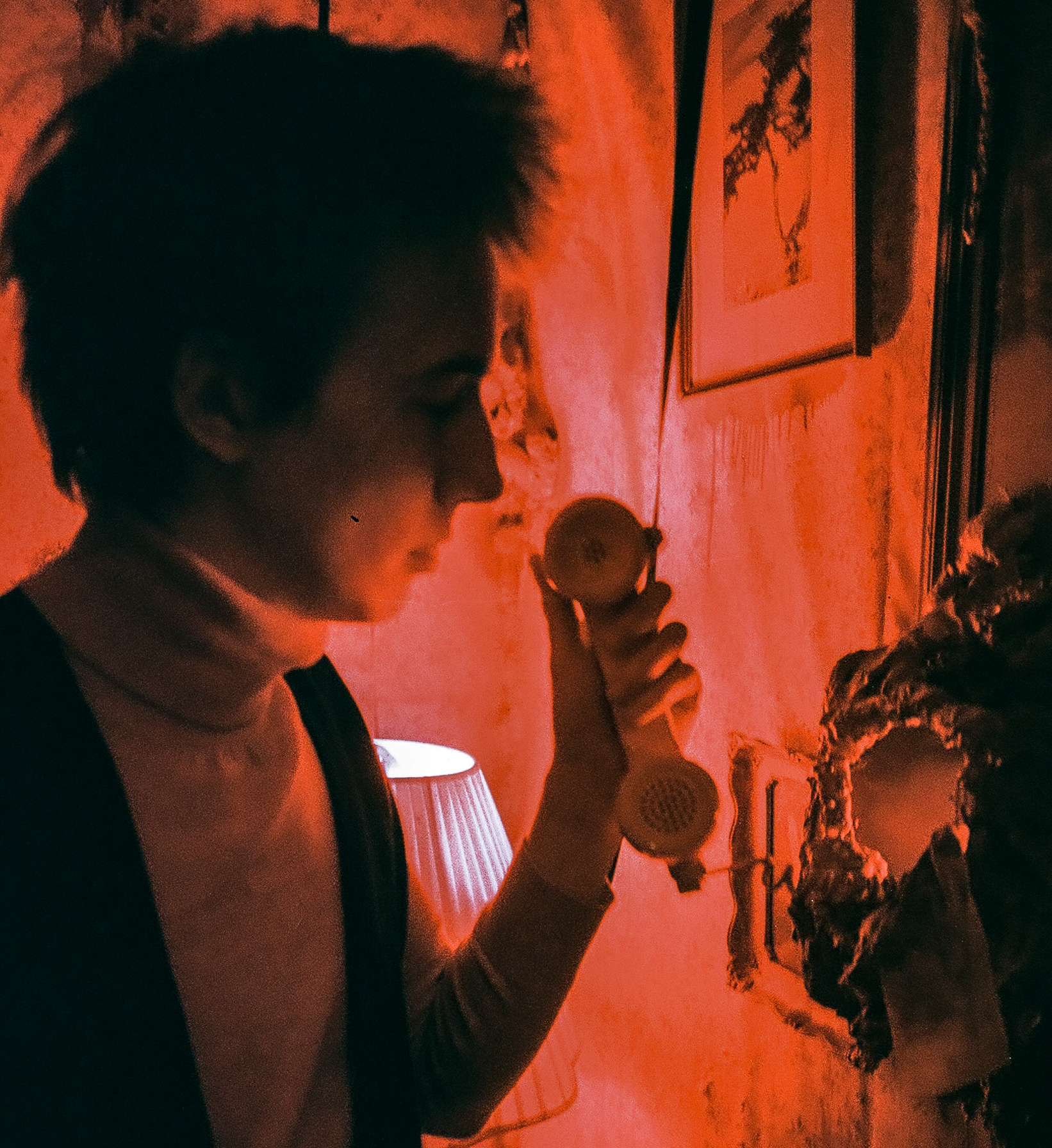 A woman with short hair is holding a vintage telephone receiver to her ear in a dimly lit room with red lighting, a lampshade, framed pictures on the wall, and a decorative mirror.