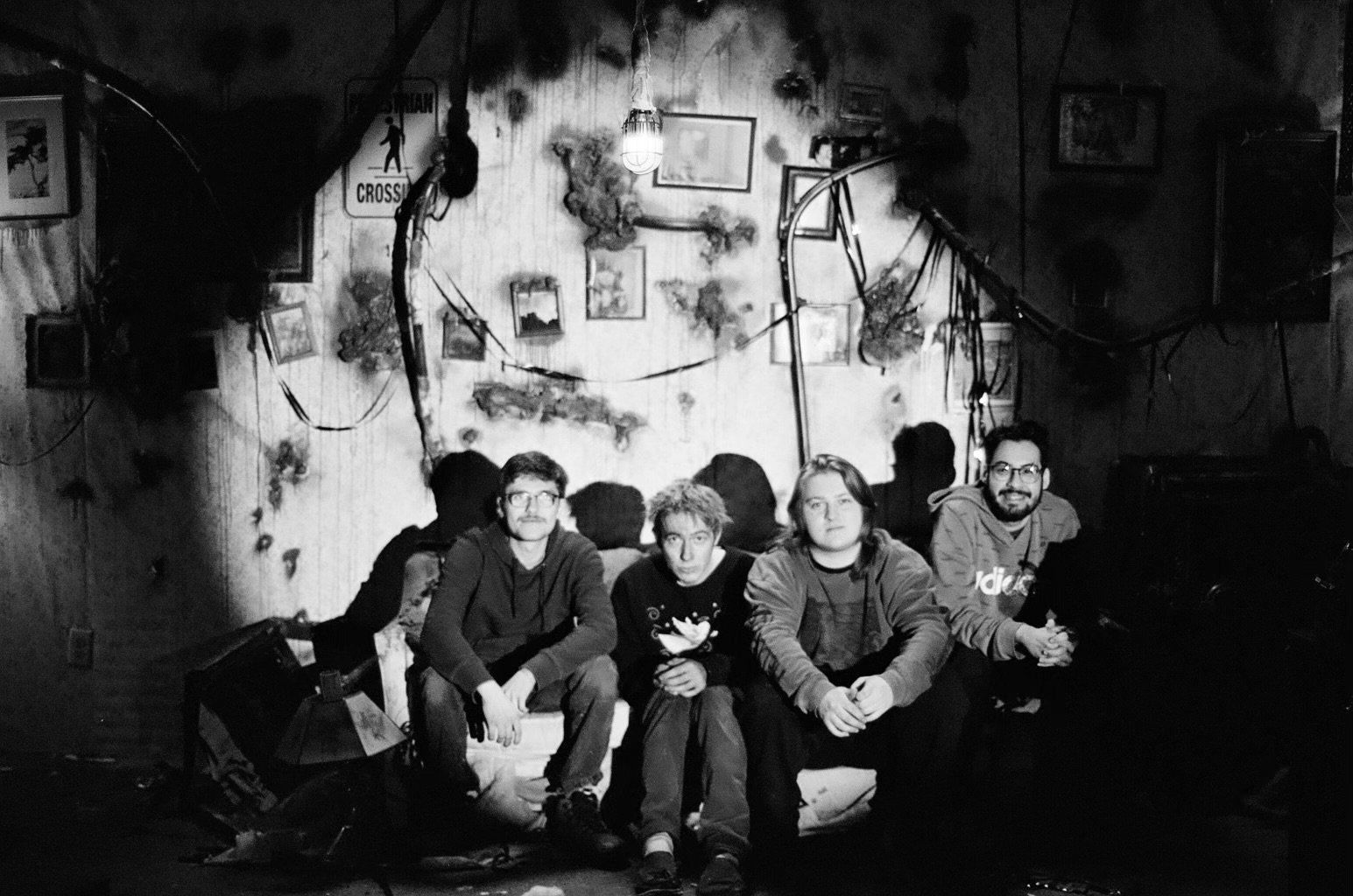 Four young men sitting on a couch in a dimly lit room with a dark and cluttered background, including pictures on the wall, a hanging light bulb, and various cables and objects.