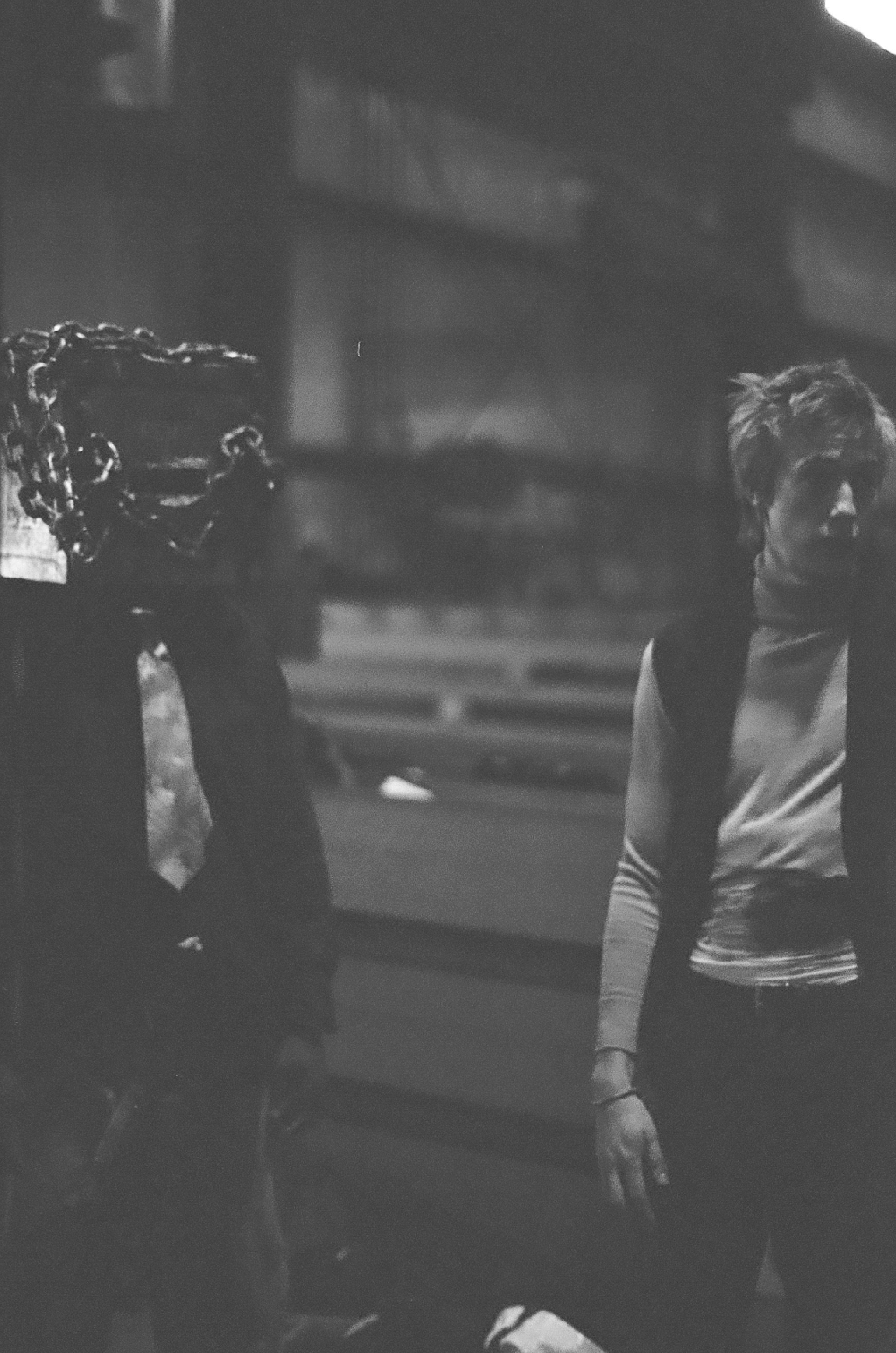 Black and white photo of two people standing indoors, with bookshelves in the background. One person is on the left, wearing a suit and tie, and the other is on the right, wearing a long-sleeve shirt, with short, tousled hair.