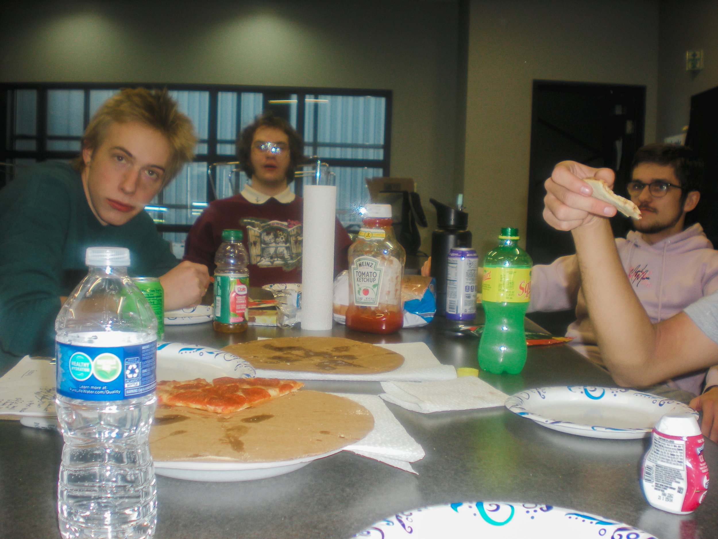 Group of four young men sitting around table with pizza, drinks, and condiments, indoors with large windows in the background.