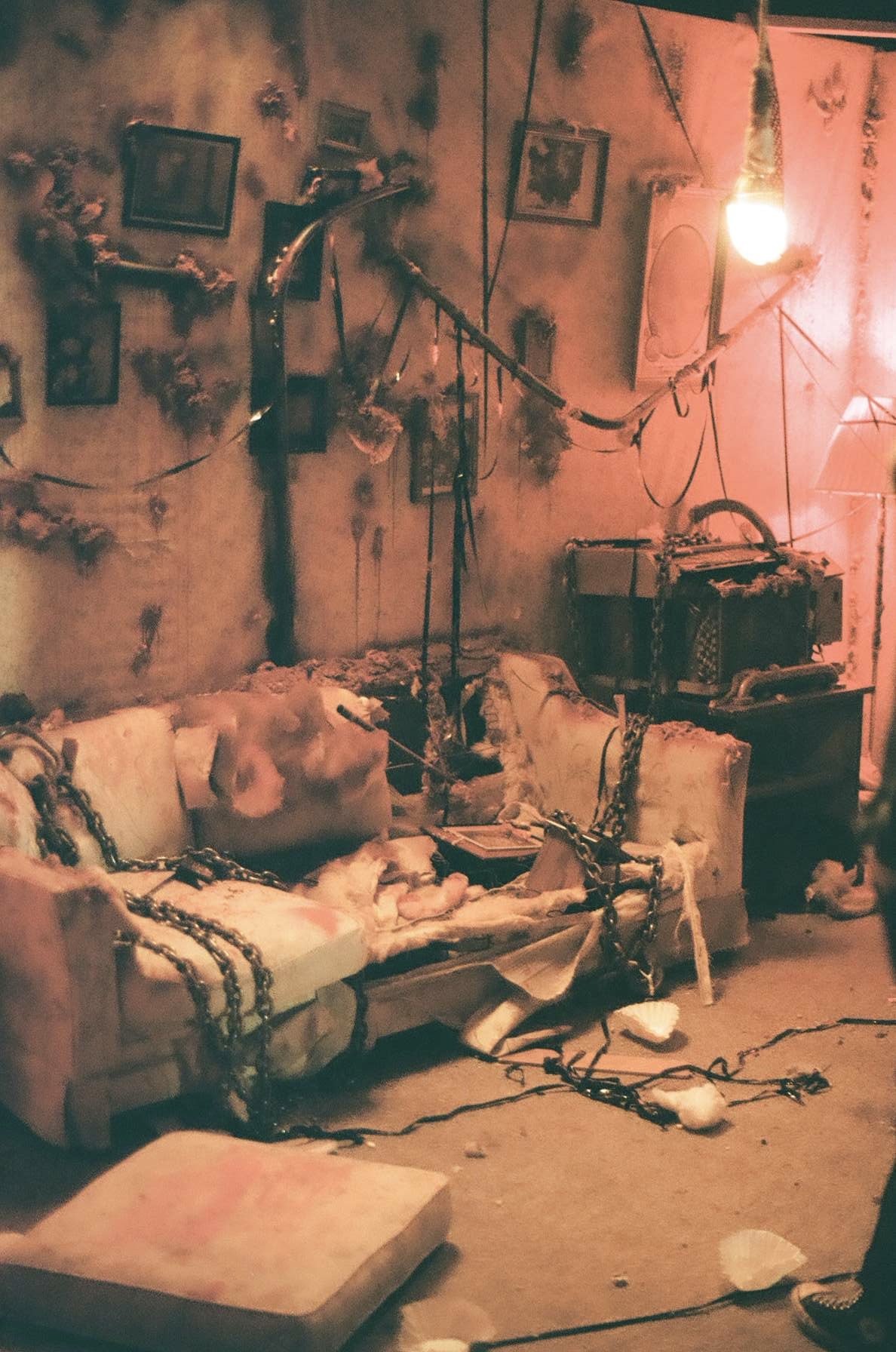 An abandoned, dilapidated living room with torn furniture, chains, and debris scattered across the floor, with a dusty, peeling wall decorated with framed pictures and a nearby exposed light bulb.