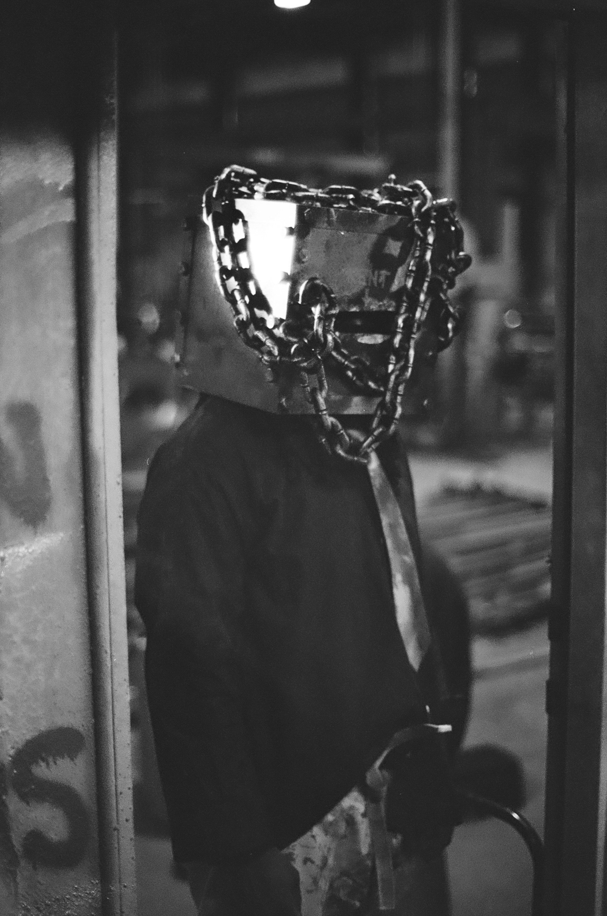 A person wearing a box over their head, which is decorated to look like a face, with chains around the box and a metal mask or face covering. They are in a dark environment with a blurred background.