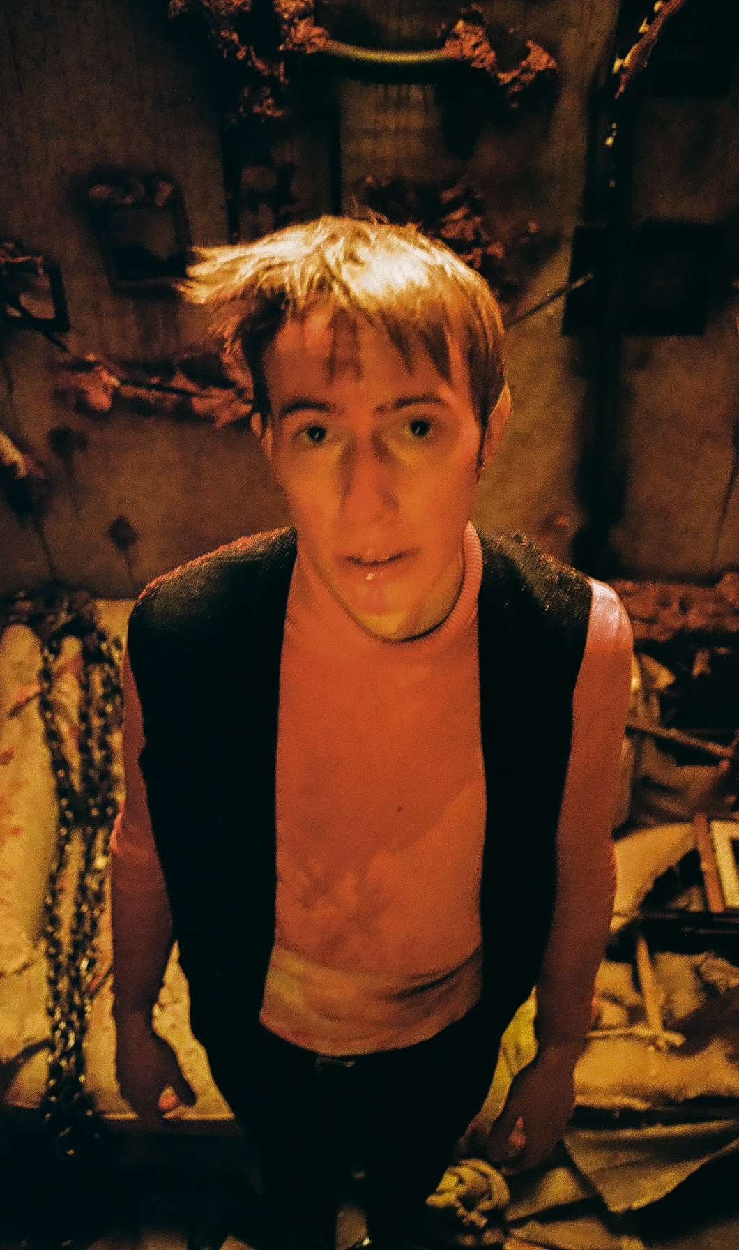 A young man with light brown hair standing in a dimly lit room with a surprised expression, with some decorations and objects on the walls behind him.