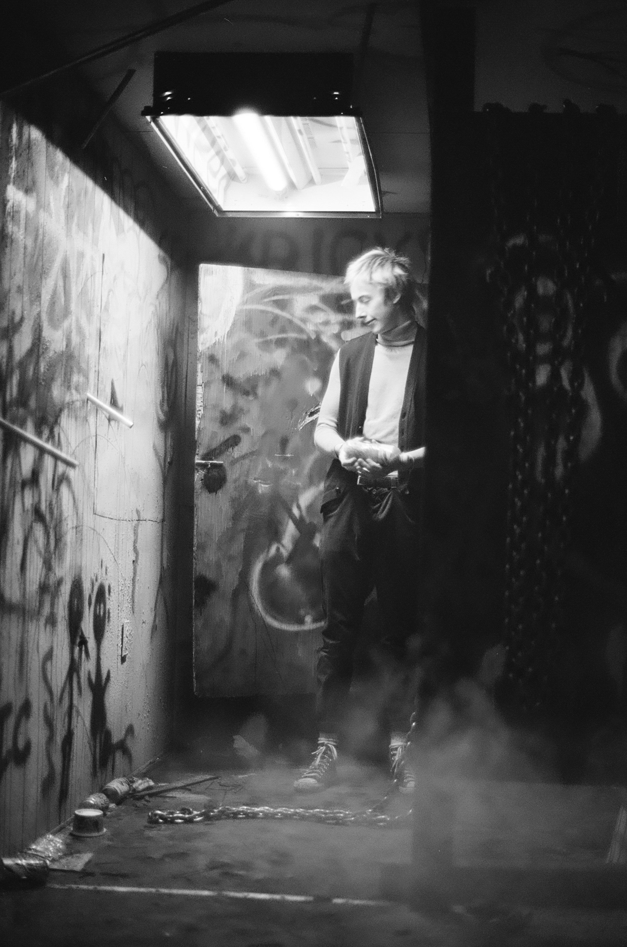 A person with short, light-colored hair standing in a dimly lit, graffiti-covered alleyway, smirking and looking down, with chains on the ground and smoke at their feet.