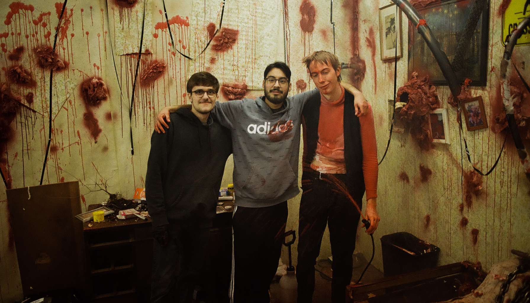 Three young men standing together in a horror-themed room covered in fake blood and gore; each has their arm around the other's shoulders. The room has messy tables and pictures on the wall.