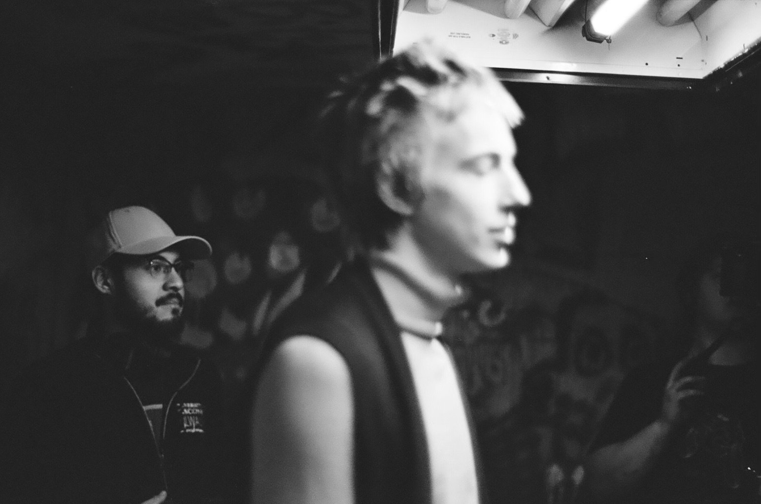 A black-and-white photo of two young men in a dimly lit setting, with the man in the foreground in profile and the other man in the background wearing glasses and a cap.