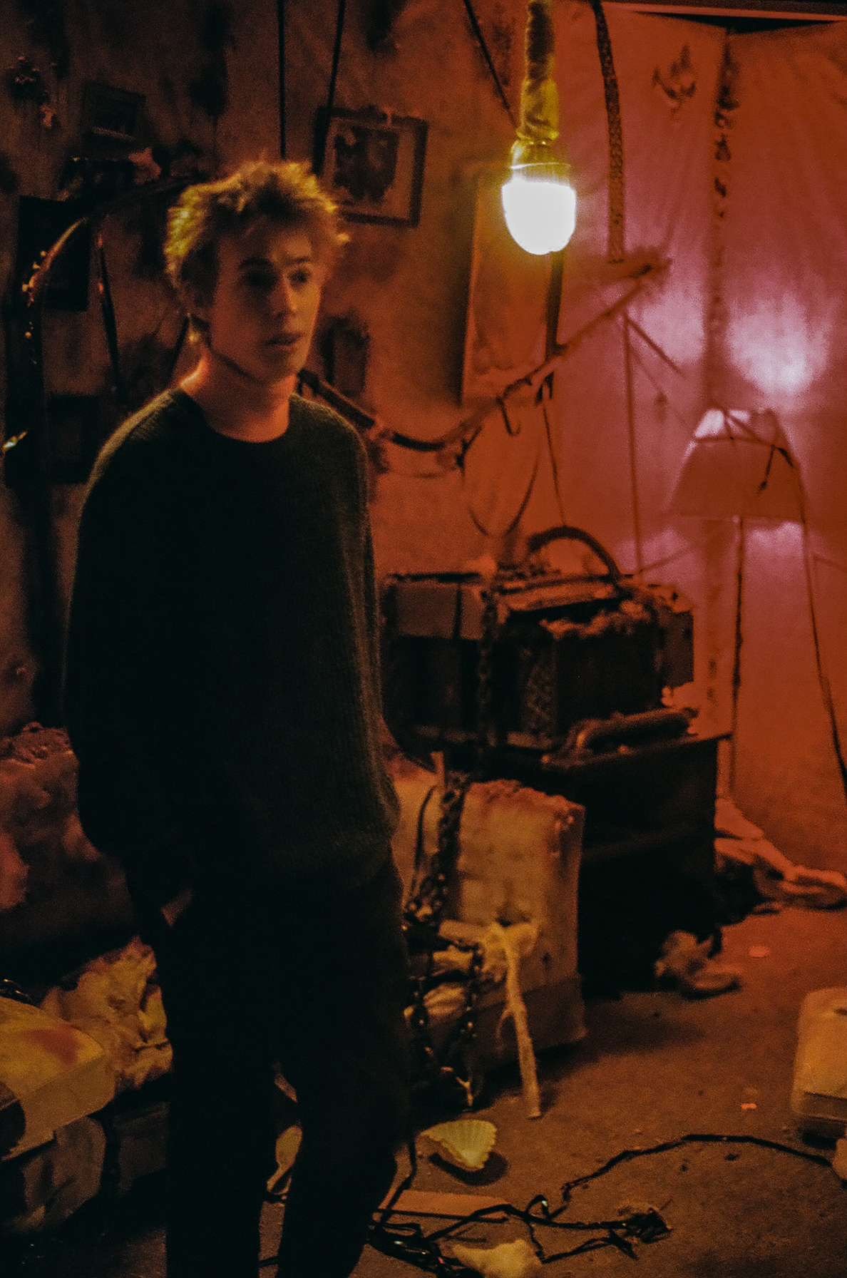A young person with messy hair stands in a dimly lit, cluttered room with pink walls, exposed wires, and a single hanging light bulb. The room has a chaotic appearance with scattered objects and furniture.