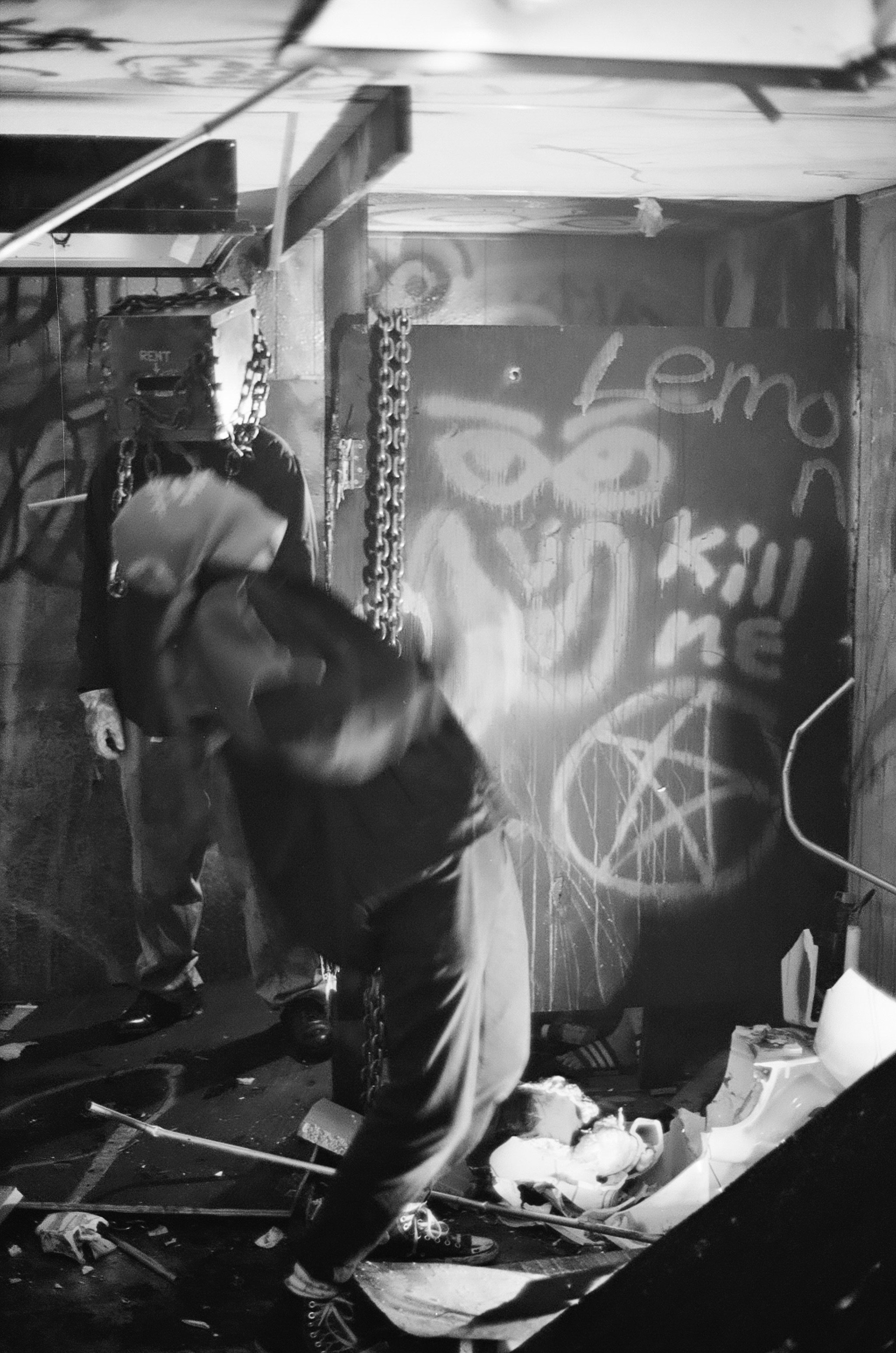A person with a chain around their neck in a room with graffiti art, including a owl and the words 'Lem' and 'Kill me,' and broken items on the floor.