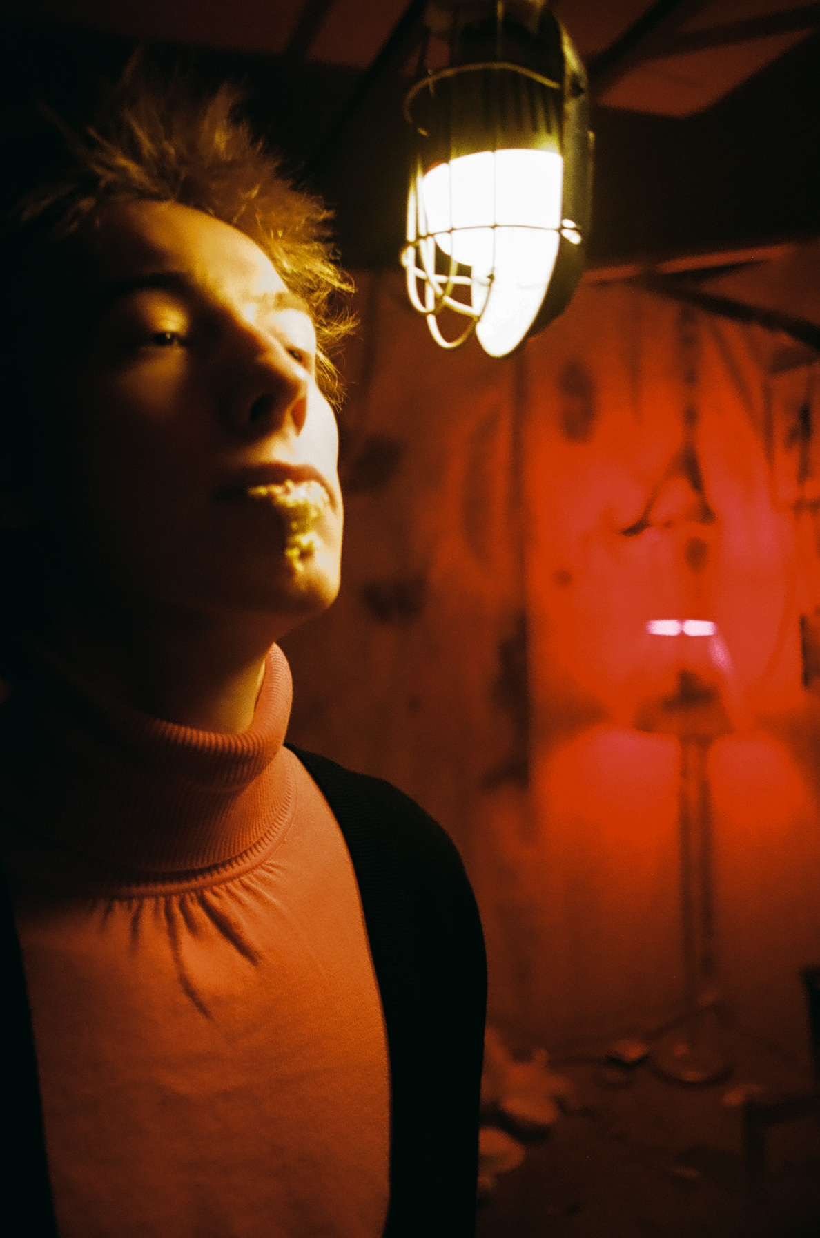 A person with short, spiky hair stands near a bright light fixture, with warm, red-toned lighting in the background, creating a moody, dramatic atmosphere.