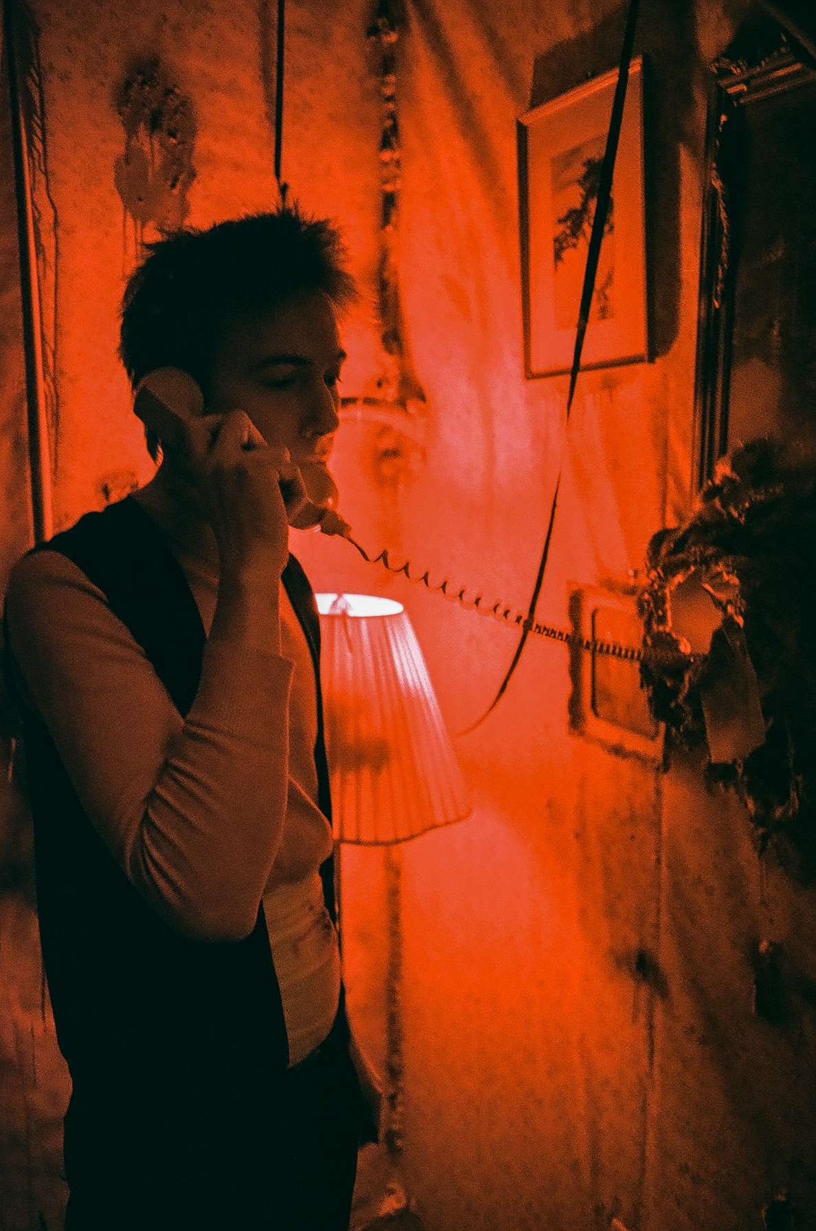 A young man with dark hair talking on a corded telephone in a dimly lit room with red lighting, vintage decor, framed pictures, and a table lamp.