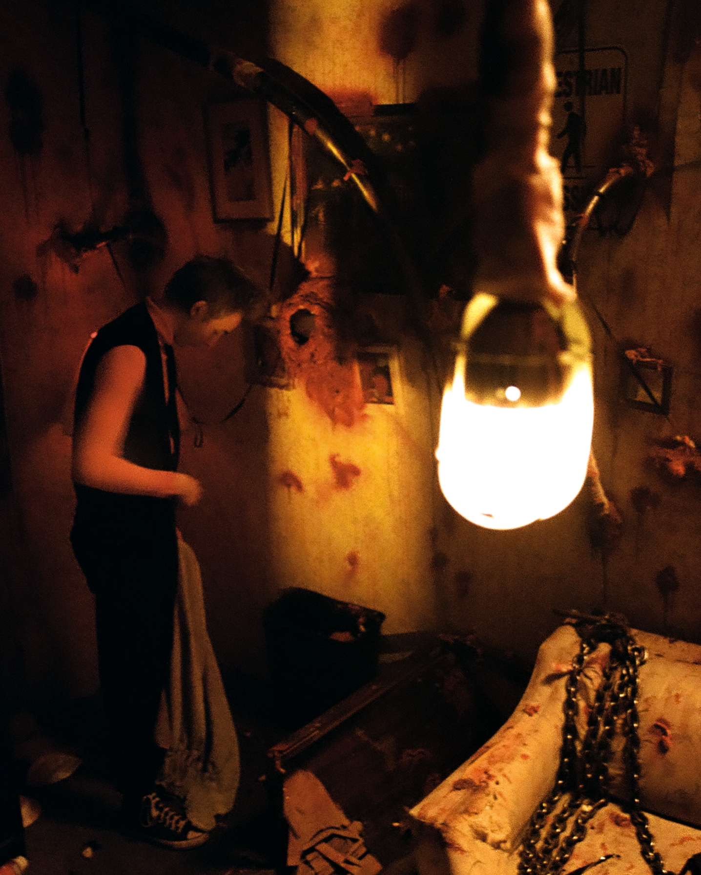 A young boy stands in a dimly lit, chaotic room with a large hanging light bulb in the foreground. The room has a worn, damaged wall decorated with framed pictures and hanging chains, with a sofa covered in chains and debris on the floor.