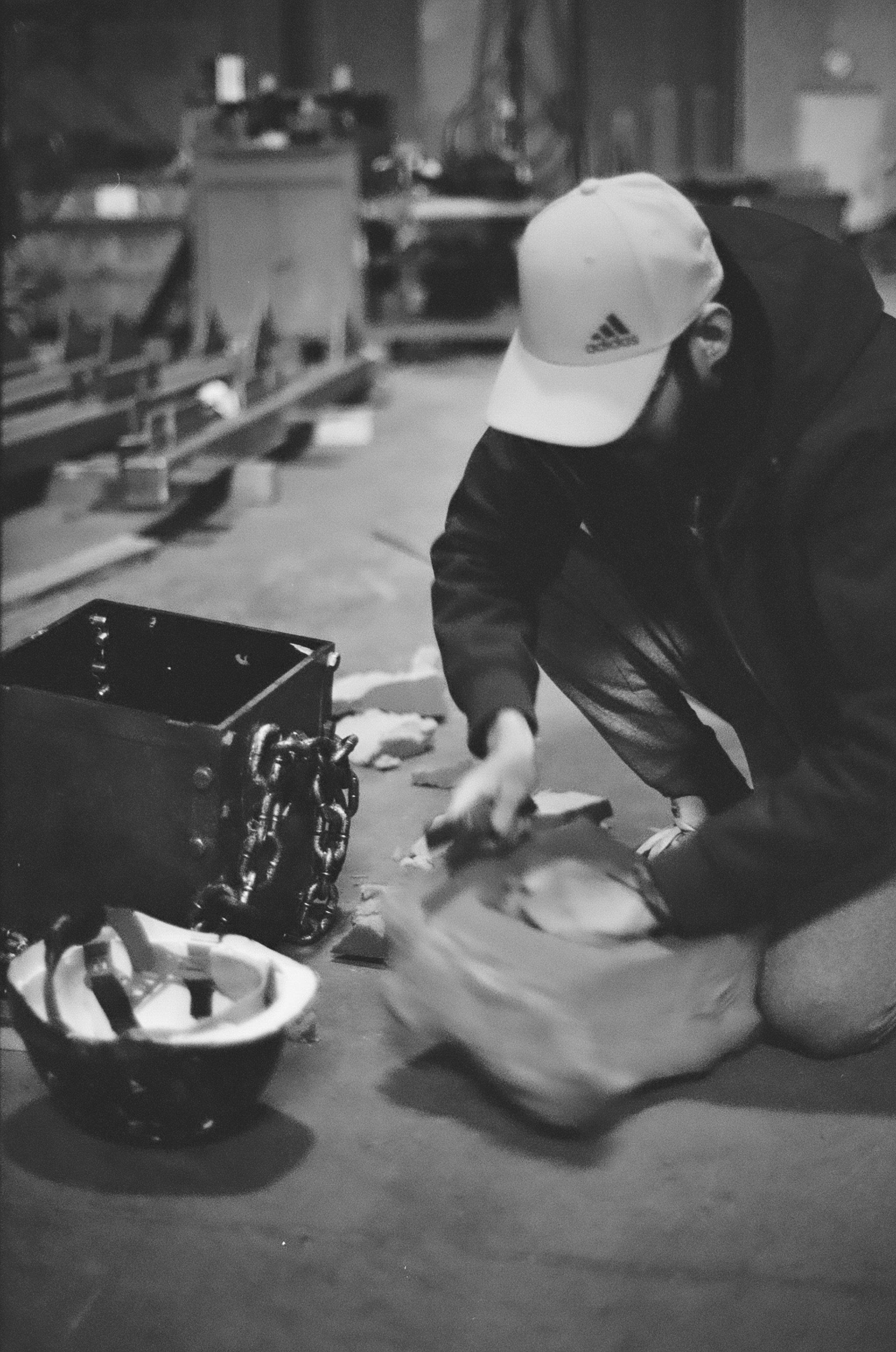A person wearing a white baseball cap and black jacket crouches down, working with a large container of fish or meat in a dimly lit indoor setting. Nearby are a helmet and a safe or box with chains.