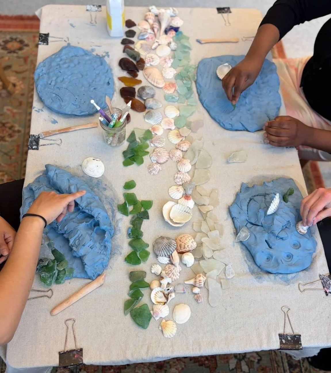 Educators engaging in a professional development Reggio-inspired provocations workshop - working on a seashell art project with various seashells and green glass pieces arranged on a table covered with a cloth.