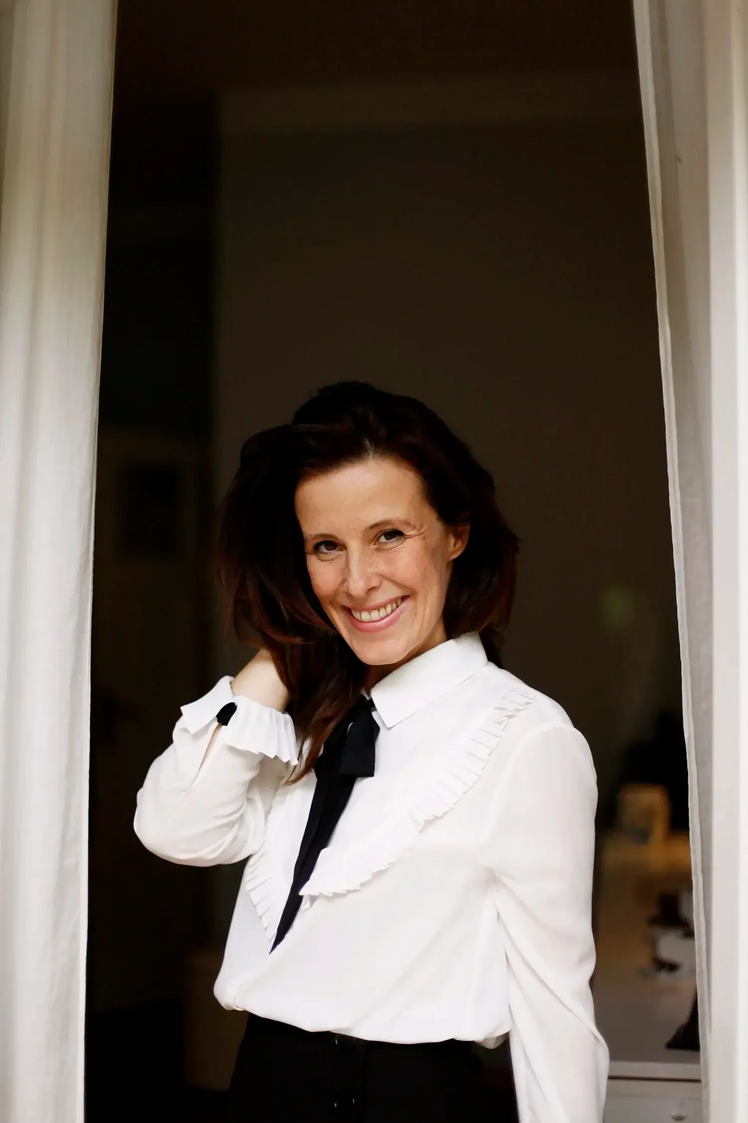 Eine Frau mit schulterlangen braunen Haaren, lächelnd, bekleidet mit einer weißen Bluse mit Rüschen und einem schwarzen Band, die in einer Tür steht.
