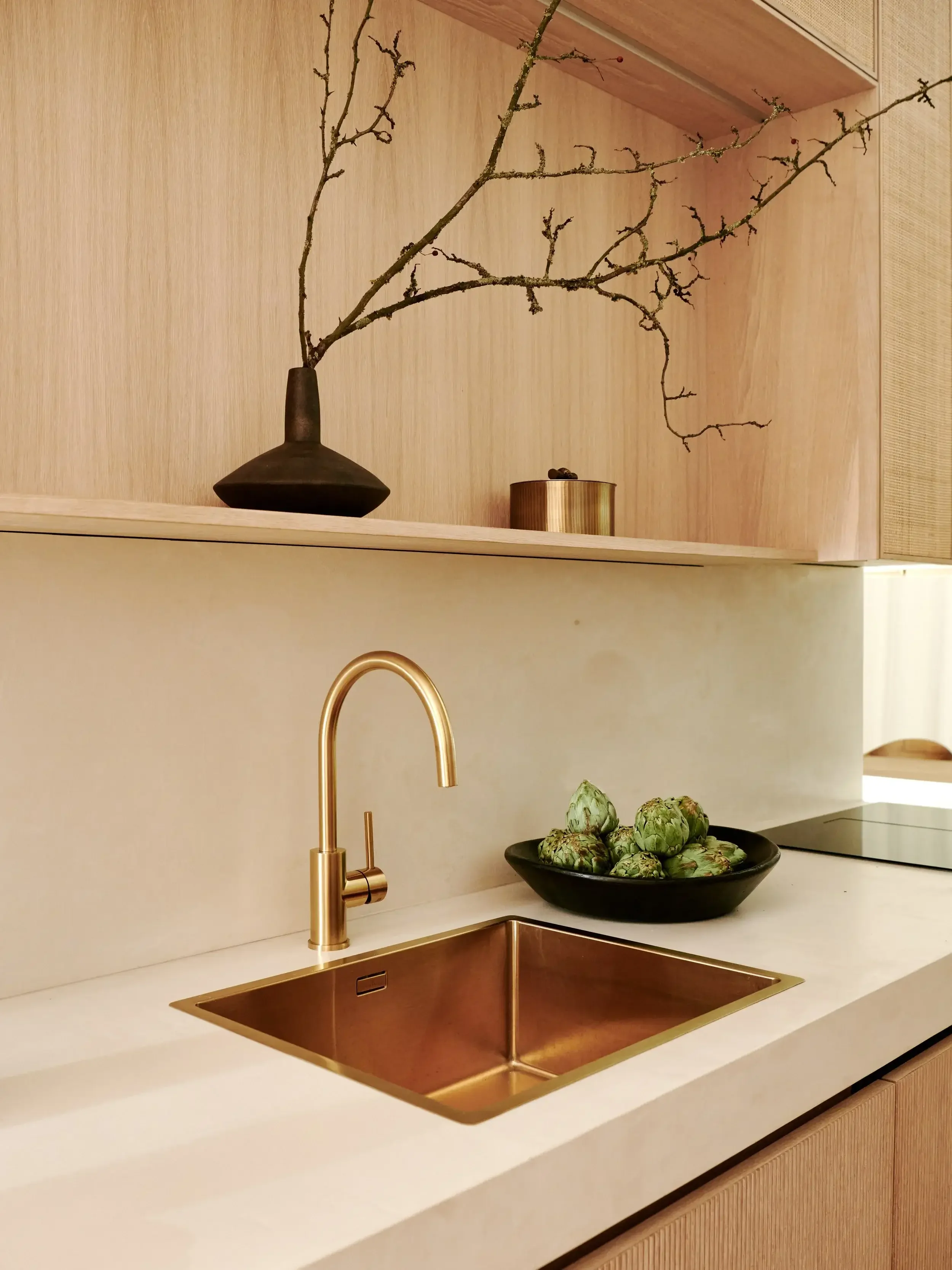 Modern kitchen with a white countertop, a gold faucet, a copper sink, a black bowl filled with artichokes, a wooden upper cabinet, and minimalist decor including a black vase with bare branches and a gold container.