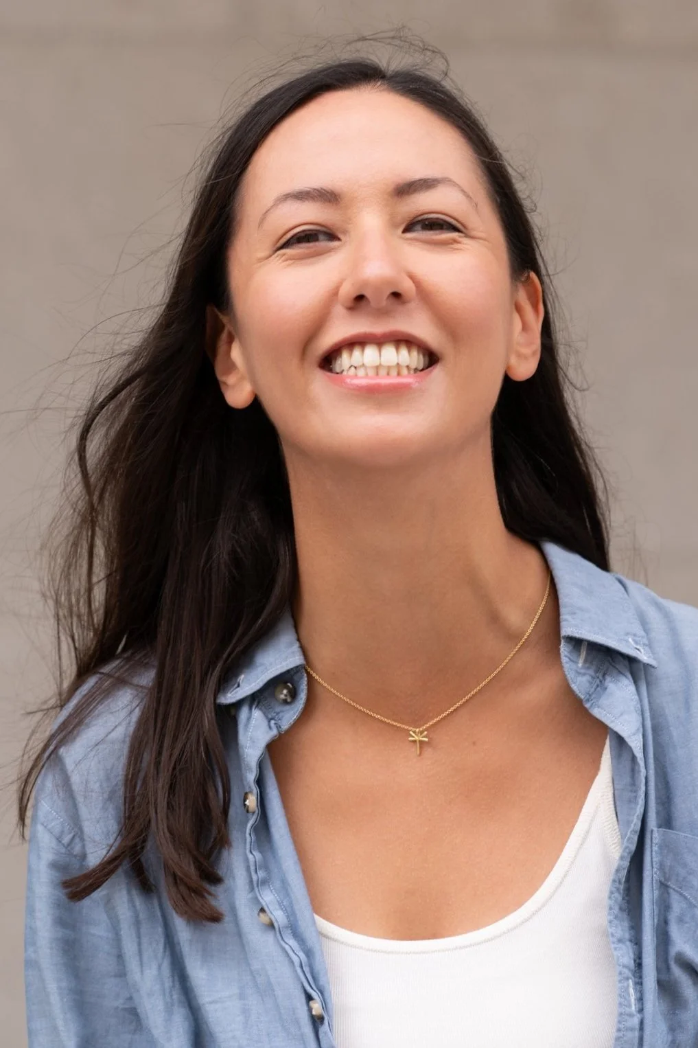 Eine Frau, die im Freien lächelt und ein Jeanshemd, ein weißes Oberteil sowie eine goldene Halskette mit einem kleinen Libellenanhänger trägt.