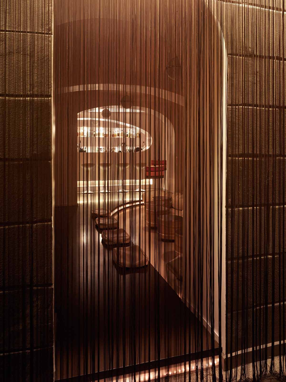 A view of a modern, illuminated bar seen through a curtain of vertical strings, with bar stools and a curved bar counter visible inside.