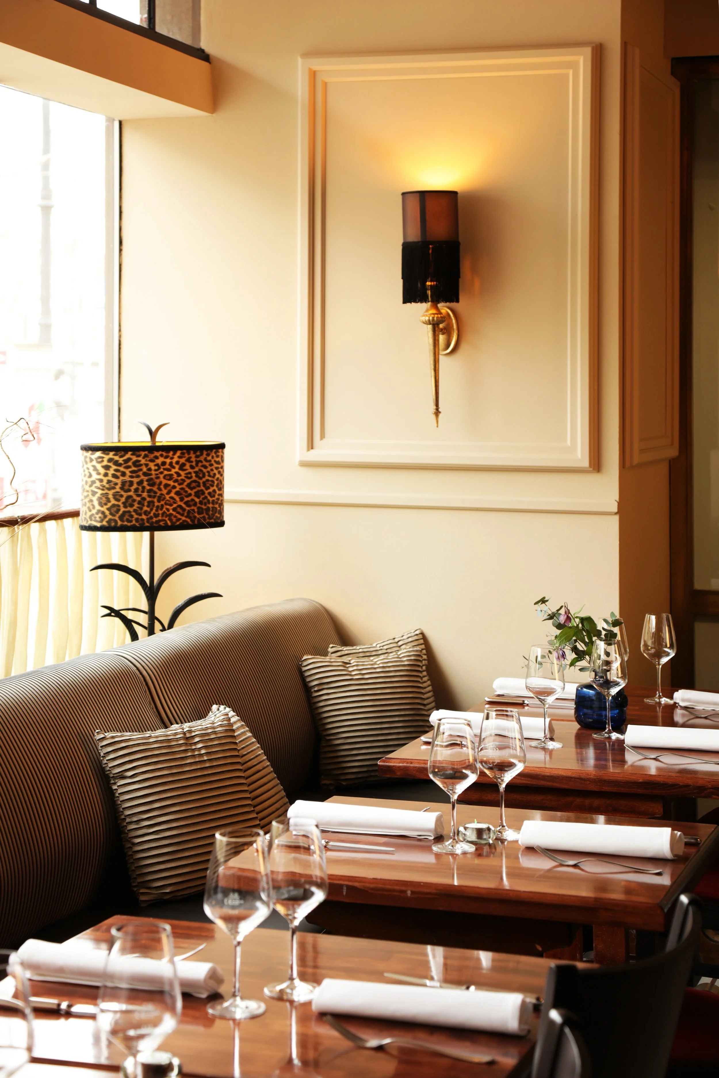 A cozy restaurant interior with a striped sofa, striped pillows, a wooden dining table set with wine glasses, napkins, and cutlery, a lamp with a leopard print lampshade, and a wall sconce. Large windows provide natural light.