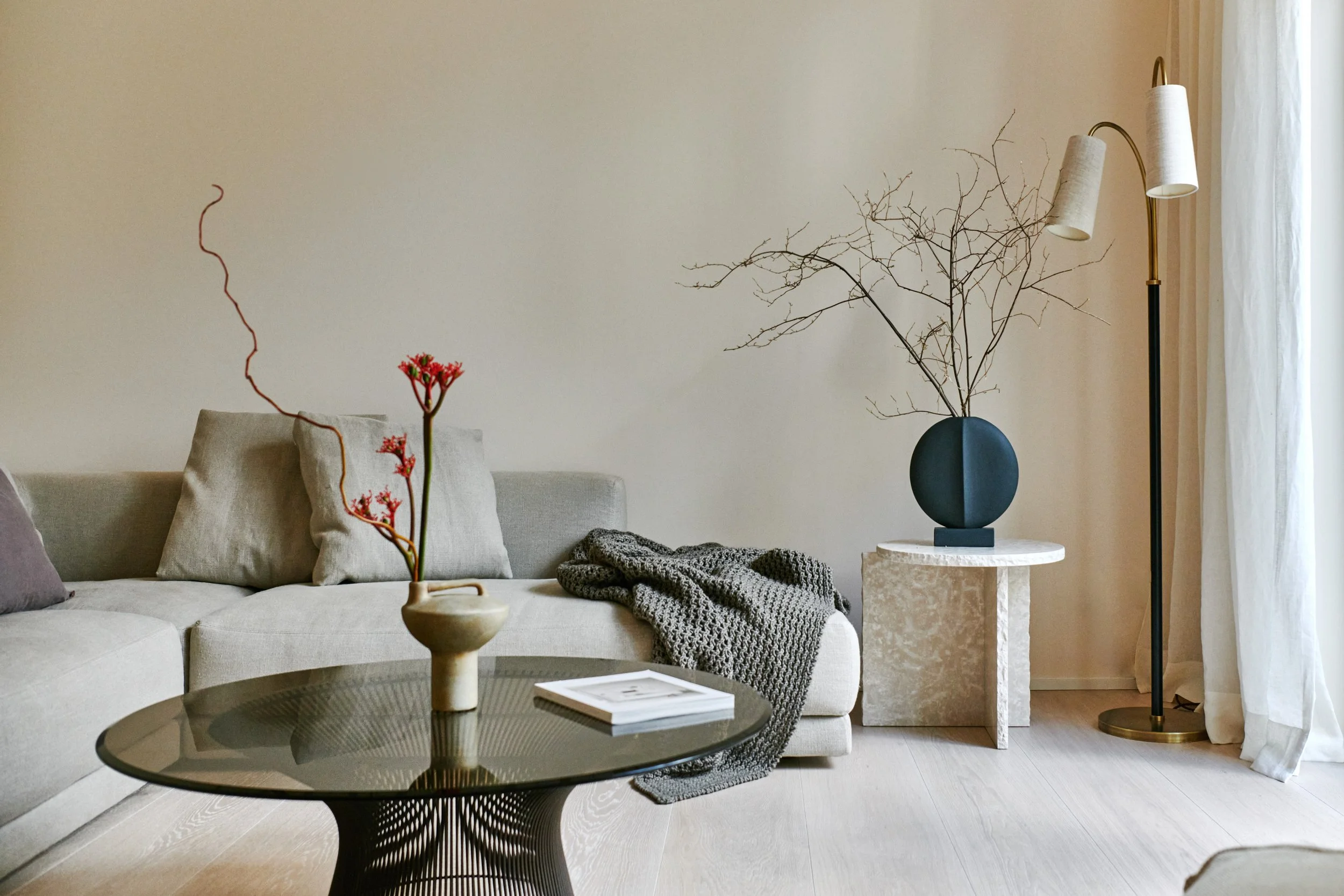 Modern living room with a beige couch, decorative pillows, a textured gray throw blanket, a black coffee table with a vase of red flowers and a book, a white marble side table with a dark blue vase holding a leafless branch, a tall black and gold floor lamp, and white curtains by a window.