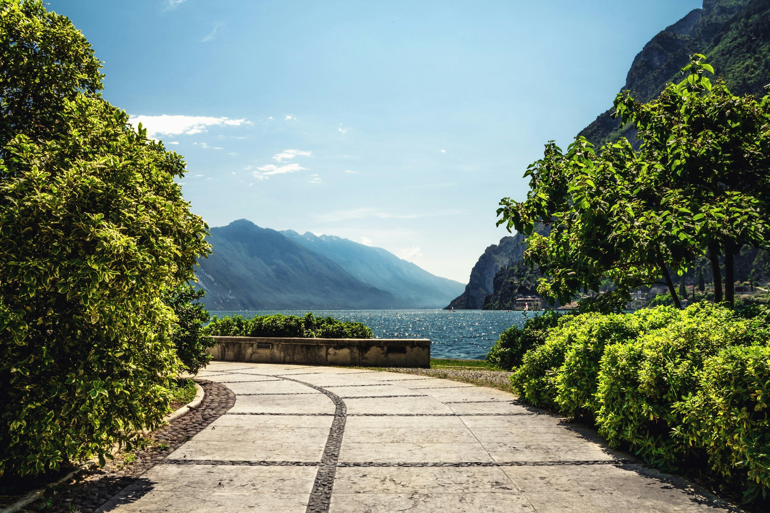 A scenic view of a lakeside with mountains in the background, framed by green bushes and trees, and a paved walkway leading to the water.
