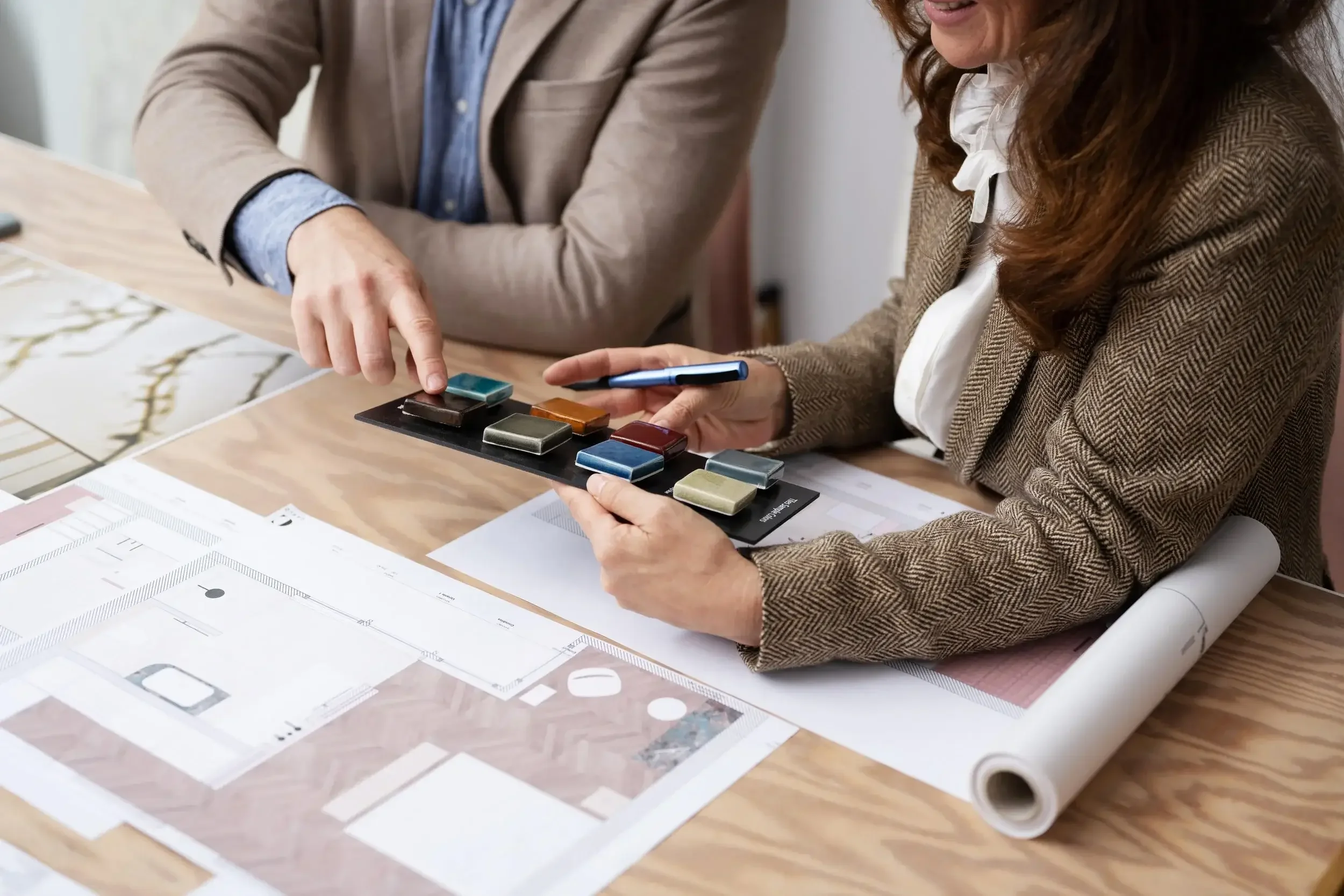 Two women dressed in business attire examining a color palette on a table, with architectural blueprints and design sketches spread out in front of them.